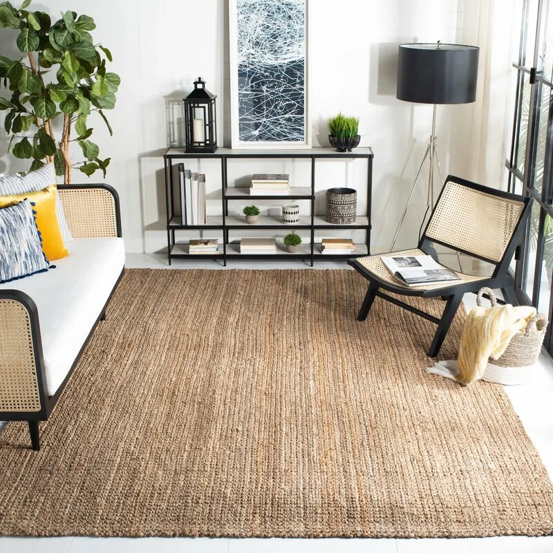 A bright living room features a white sofa with cane panels, a black cane chair, a large textured jute rug, and a metal console table.