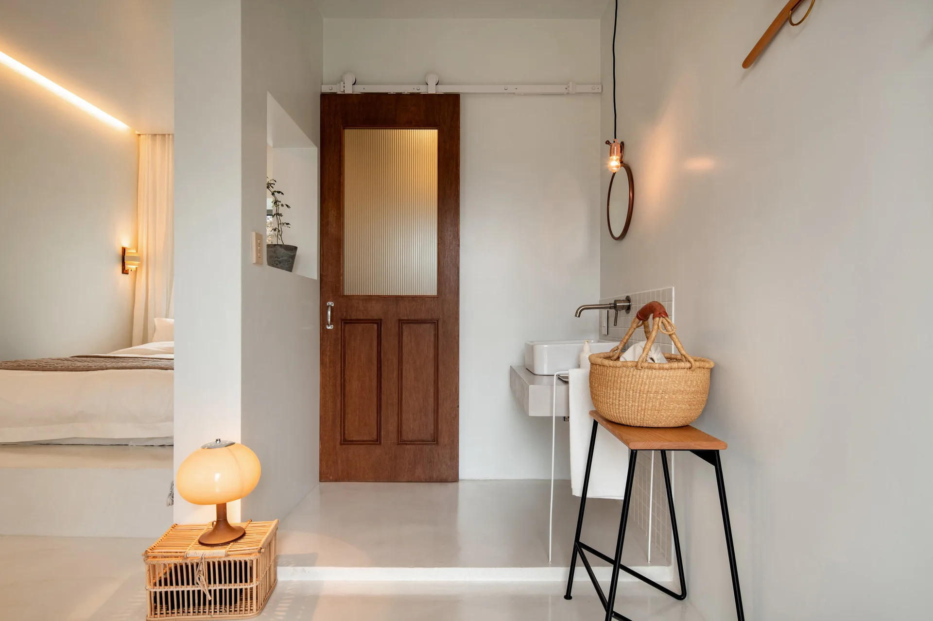 A minimalist bathroom features a dark wood sliding door with ribbed glass, a wall-mounted sink, and a raised bedroom platform in the background.