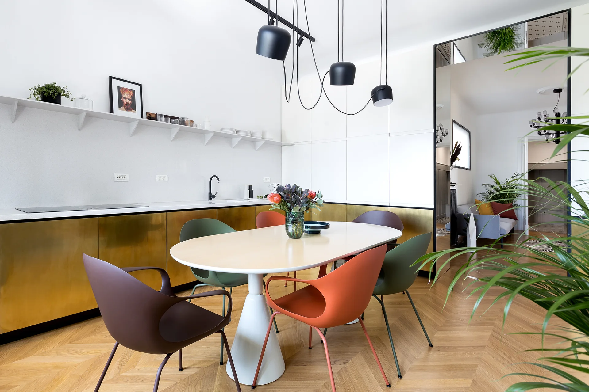 A modern kitchen features gold metallic lower cabinets, an oval dining table with colorful chairs, and draped black pendant lights over a herringbone floor.