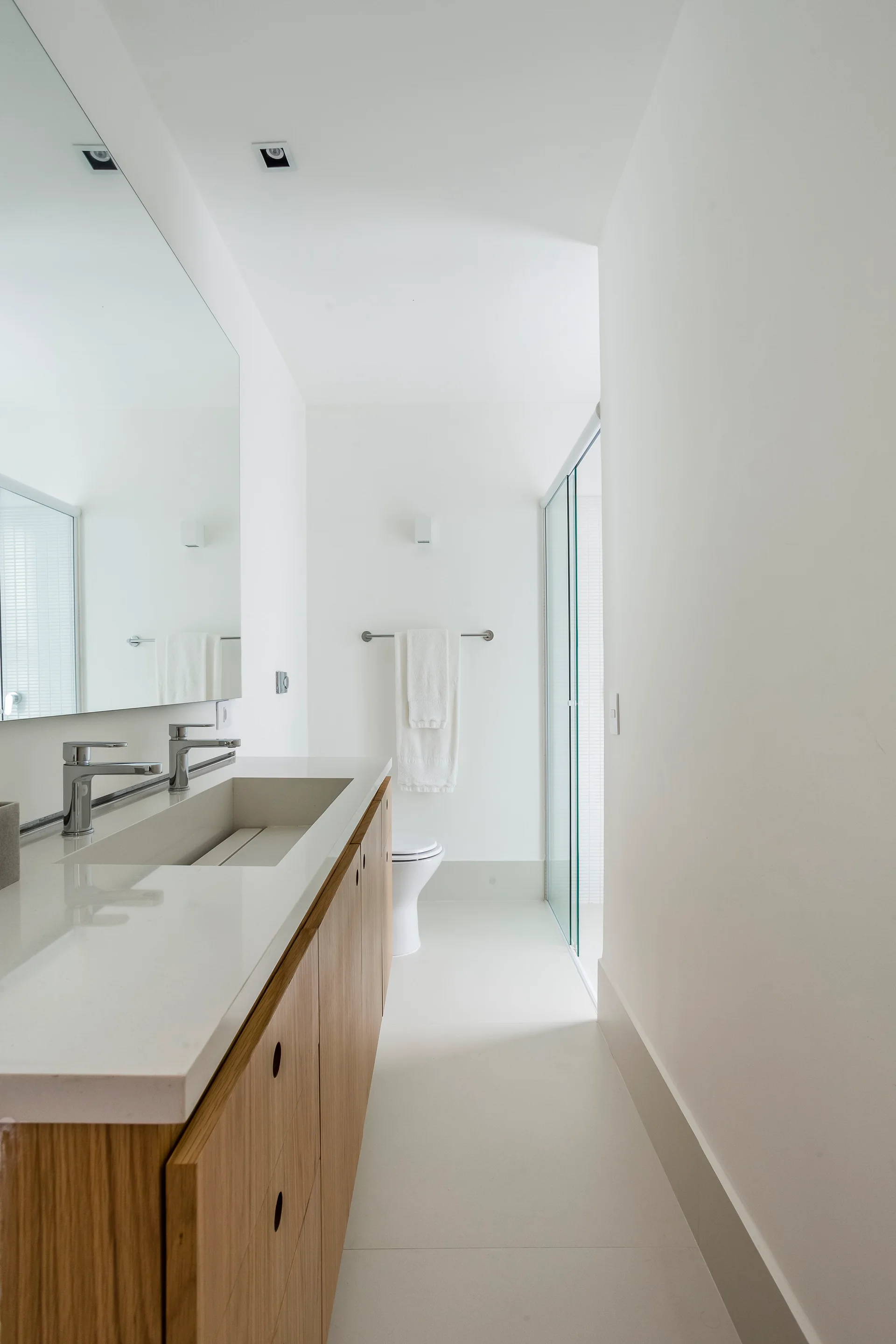 A modern white bathroom featuring a warm wood vanity with an integrated white sink, dual chrome faucets, and a large frameless mirror.