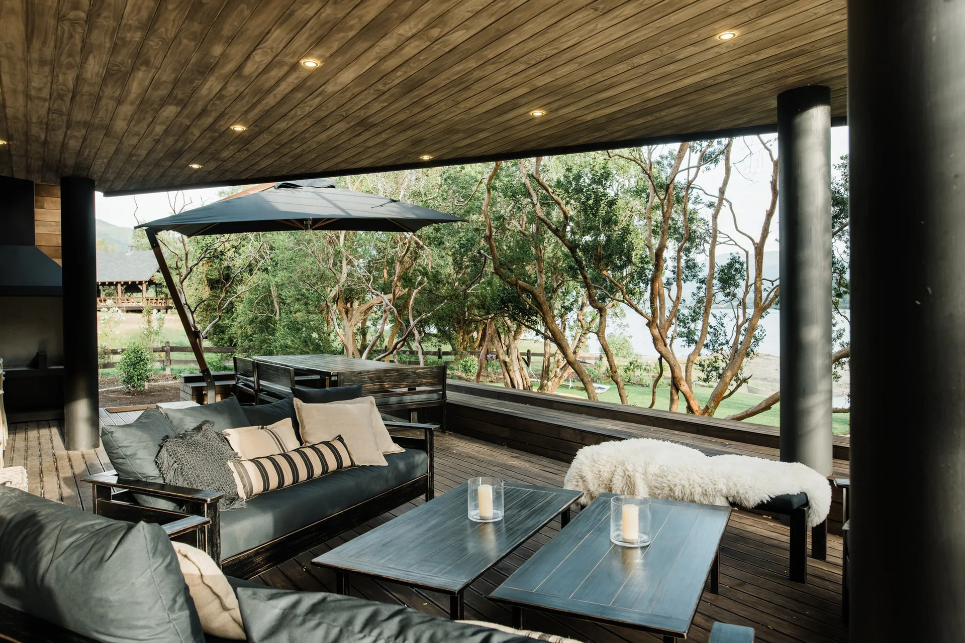 A covered wood patio features a charcoal grey sofa, twin coffee tables, and a wood plank ceiling looking out toward green trees.