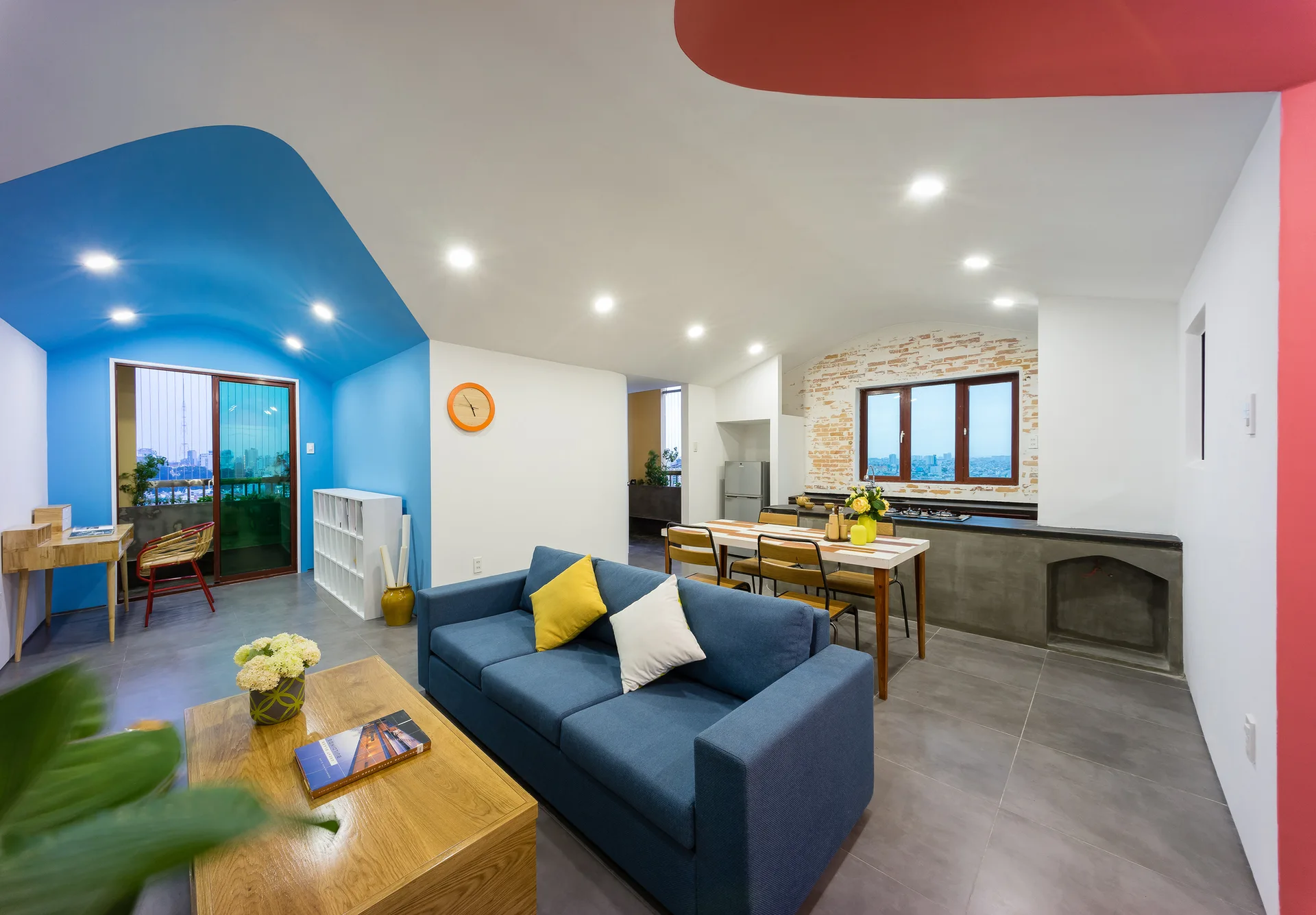 An open-plan living and dining area with a blue sofa, a wooden dining table, gray tiled floors, and vibrant blue and red color-blocked walls.