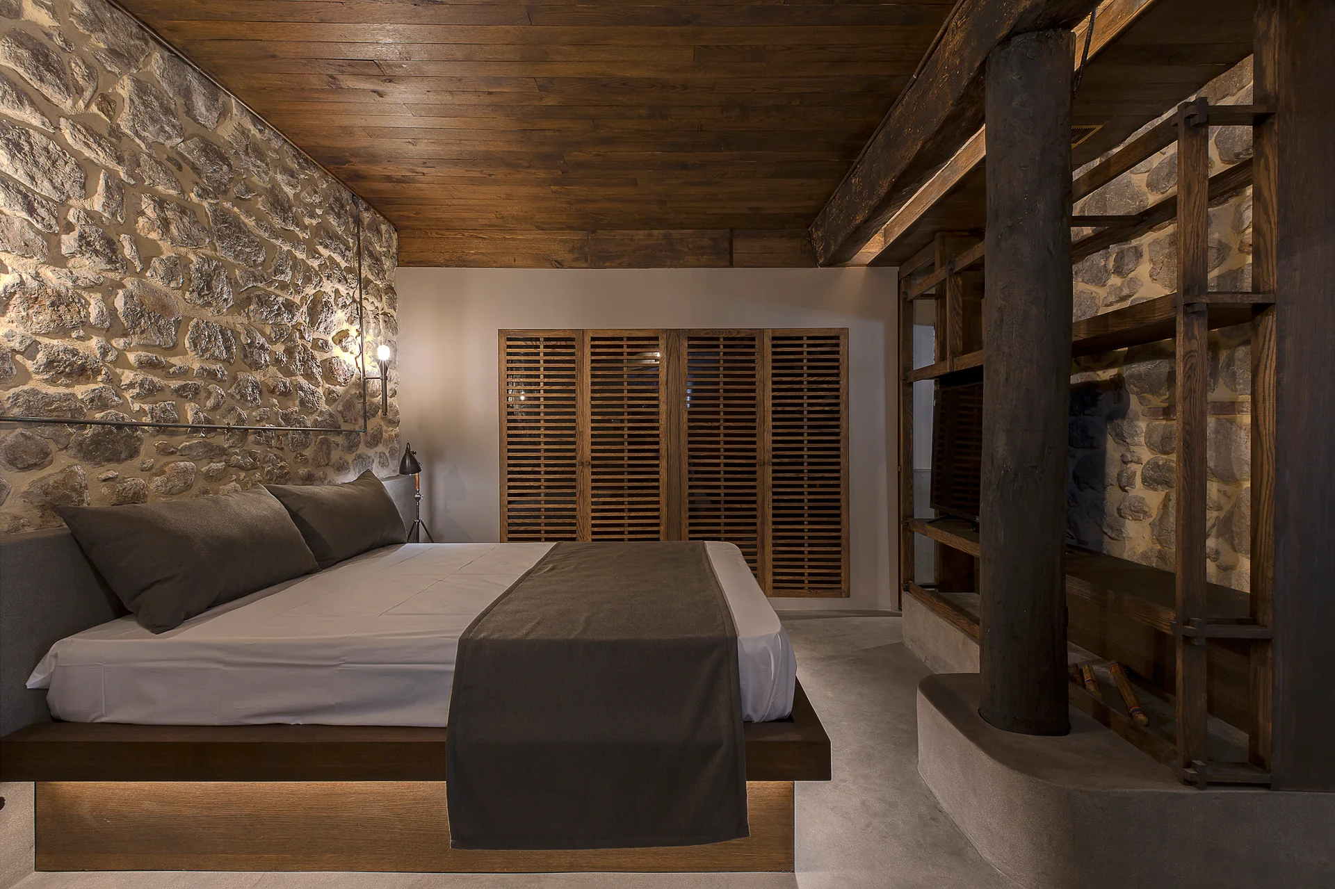 A rustic bedroom with an exposed stone accent wall, a low platform bed with under-bed lighting, wood-paneled ceilings, and louvered closet doors.