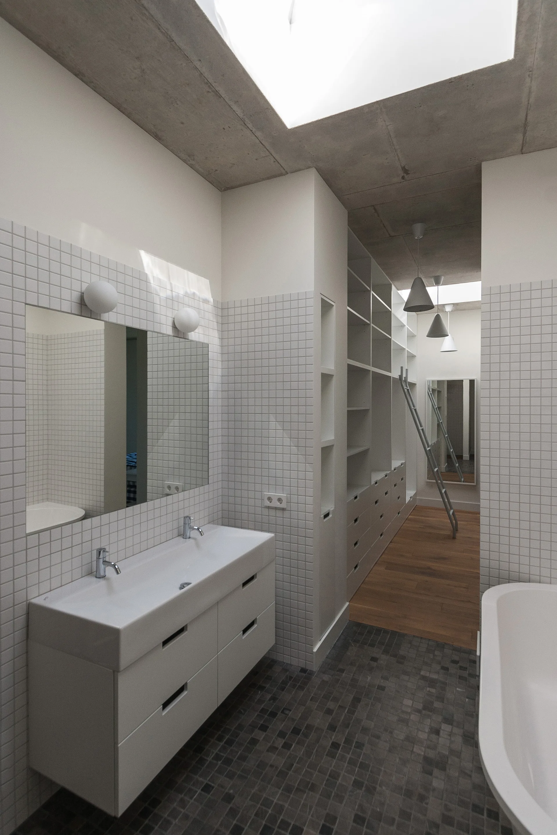 A minimalist bathroom with a white double vanity, grid tile walls, an exposed concrete ceiling, and an open dressing closet in the background.