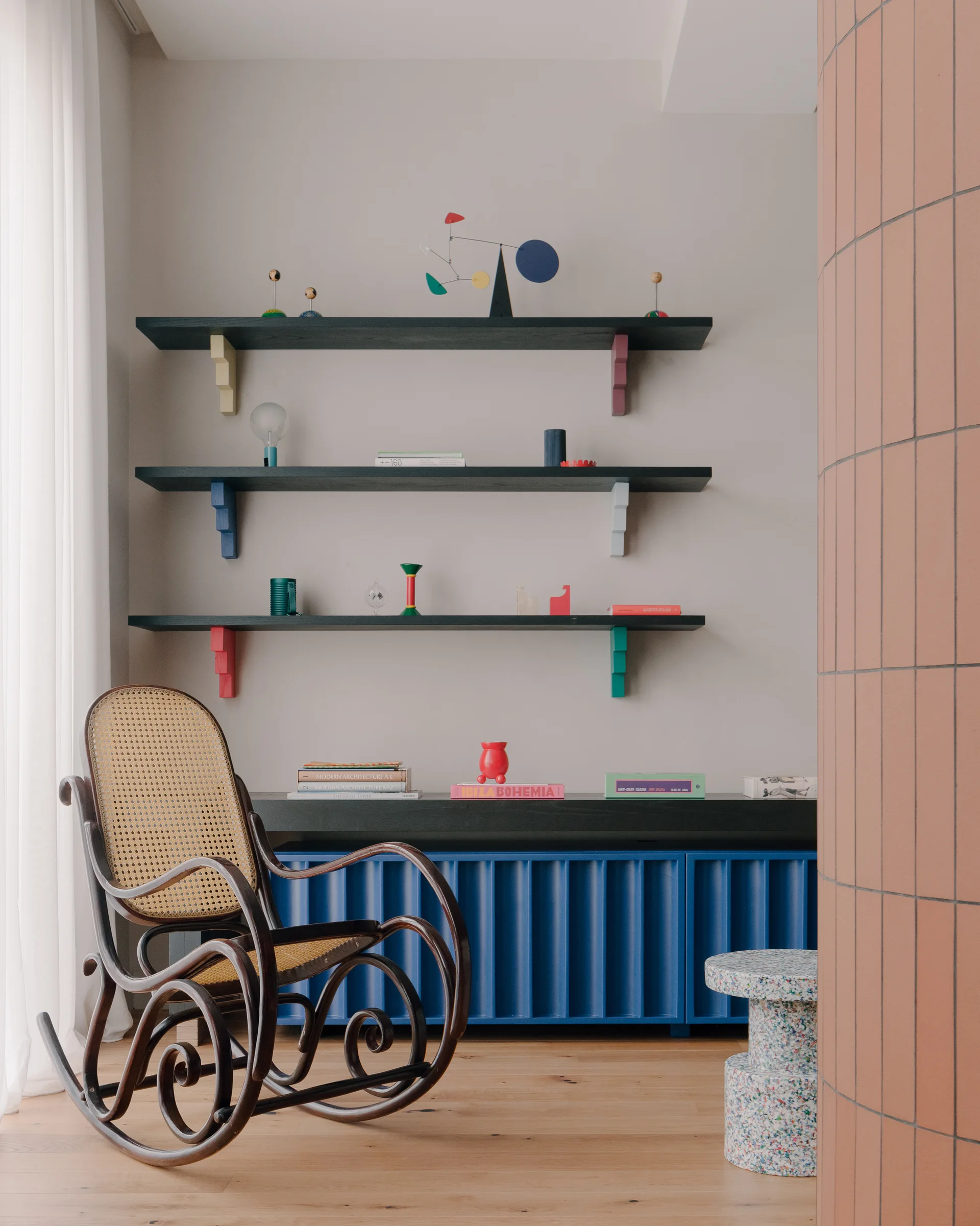 A classic bentwood rocking chair with a cane back sits next to a fluted blue console and black floating shelves against a grey wall.