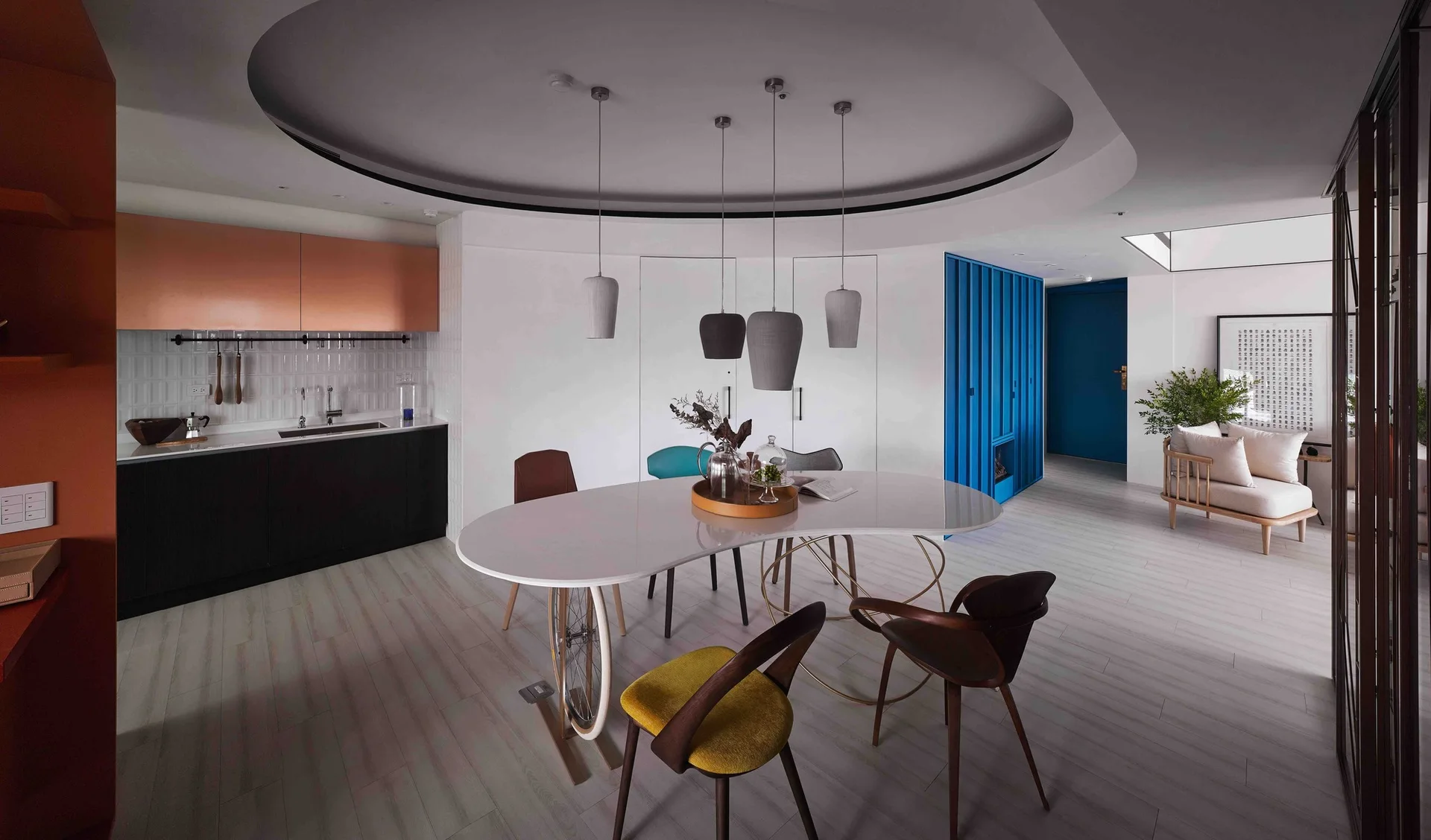 An organic-shaped white dining table with mixed-style chairs sits beneath a circular recessed ceiling next to a kitchen with copper cabinets.