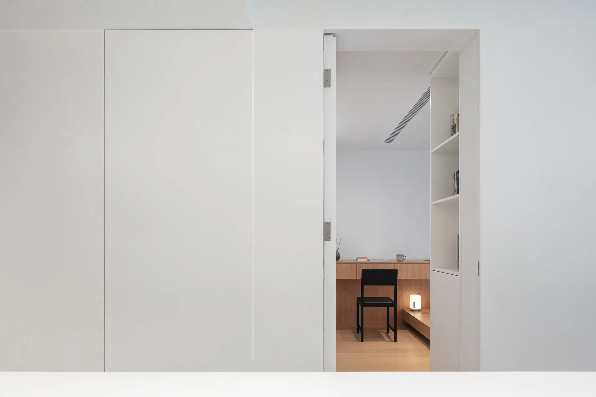 A white doorway with integrated shelving opens to a minimalist home office featuring a light wood desk, black chair, and low warm lighting.