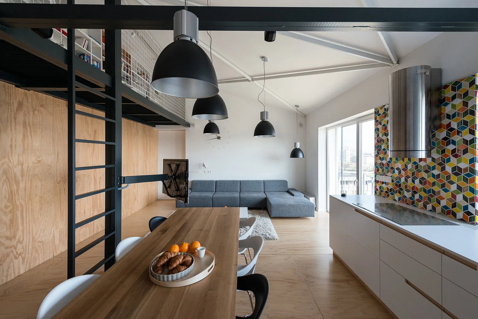 An open-plan loft apartment featuring a kitchen with geometric backsplash tiles, a long wooden dining table, a mezzanine ladder, and a grey sectional sofa.