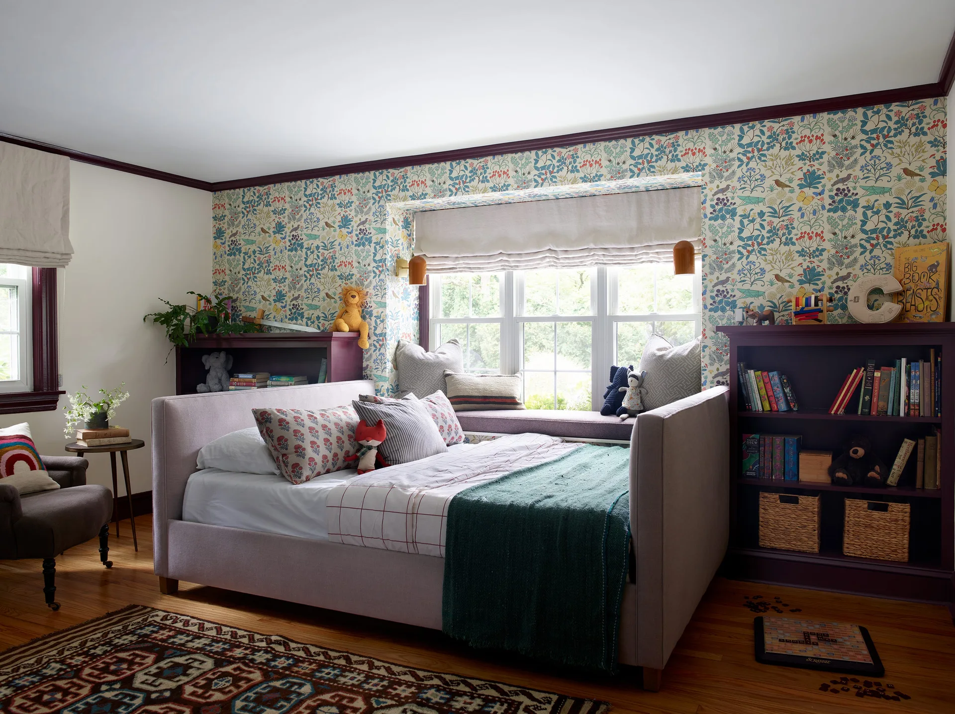 A child's bedroom featuring an upholstered daybed in front of a window seat flanked by deep purple built-in bookshelves and floral wallpaper.