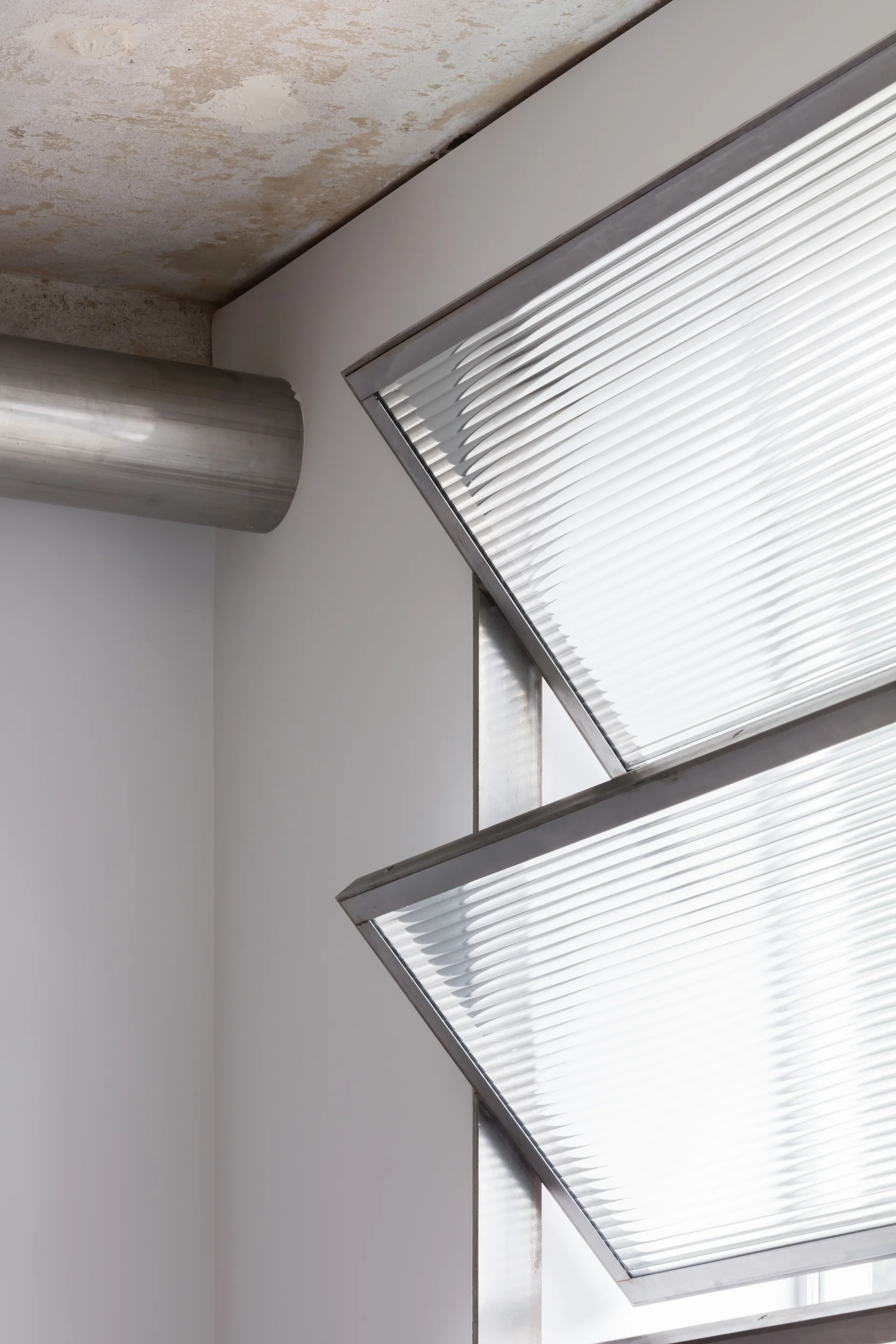 An industrial interior detail showing an open hinged window with fluted glass panes, a white wall, a metal duct, and a raw concrete ceiling.