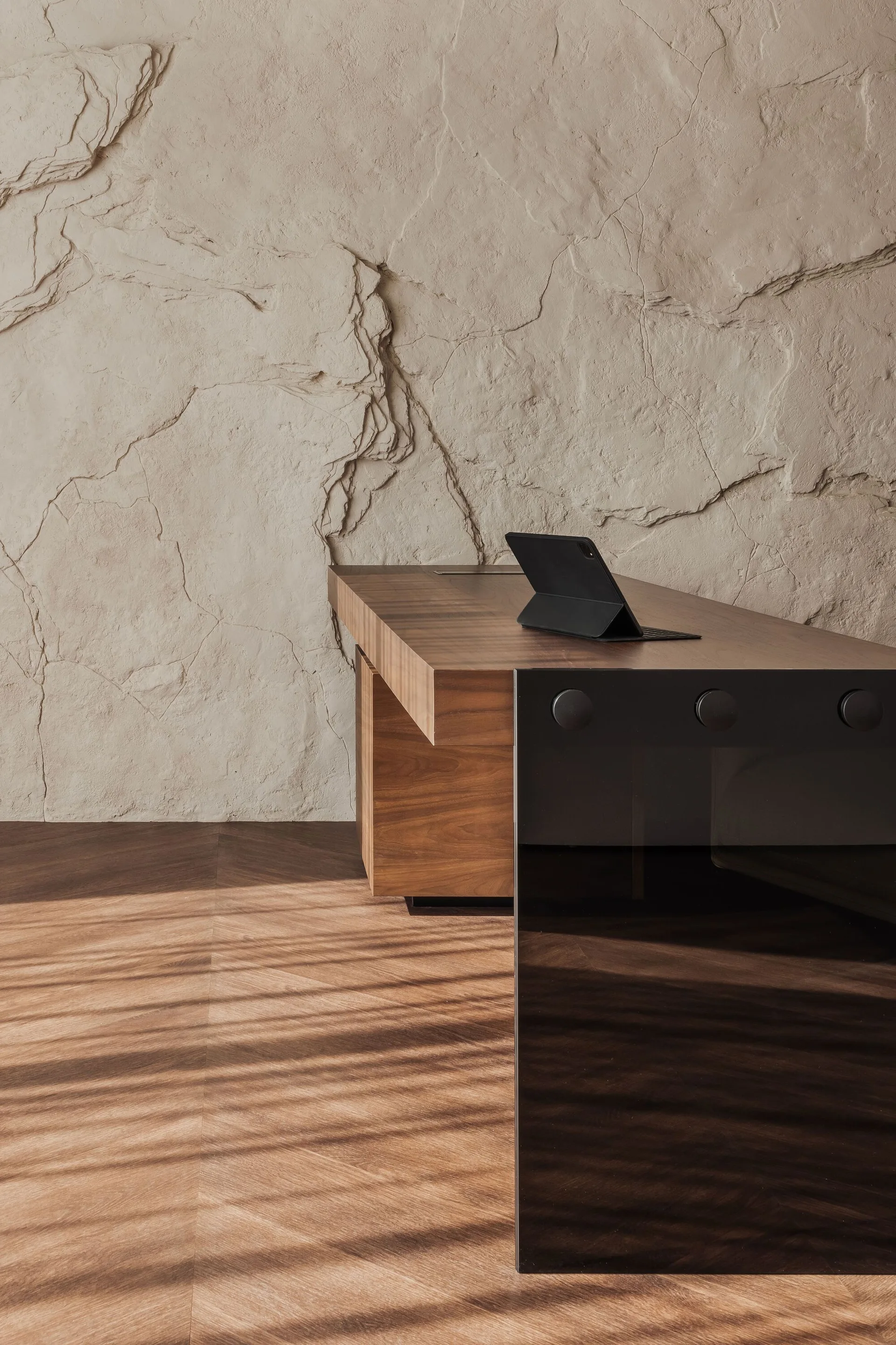 A large wooden desk with a black tablet sits on a chevron wood floor in front of a heavily textured beige stone wall with dappled sunlight.