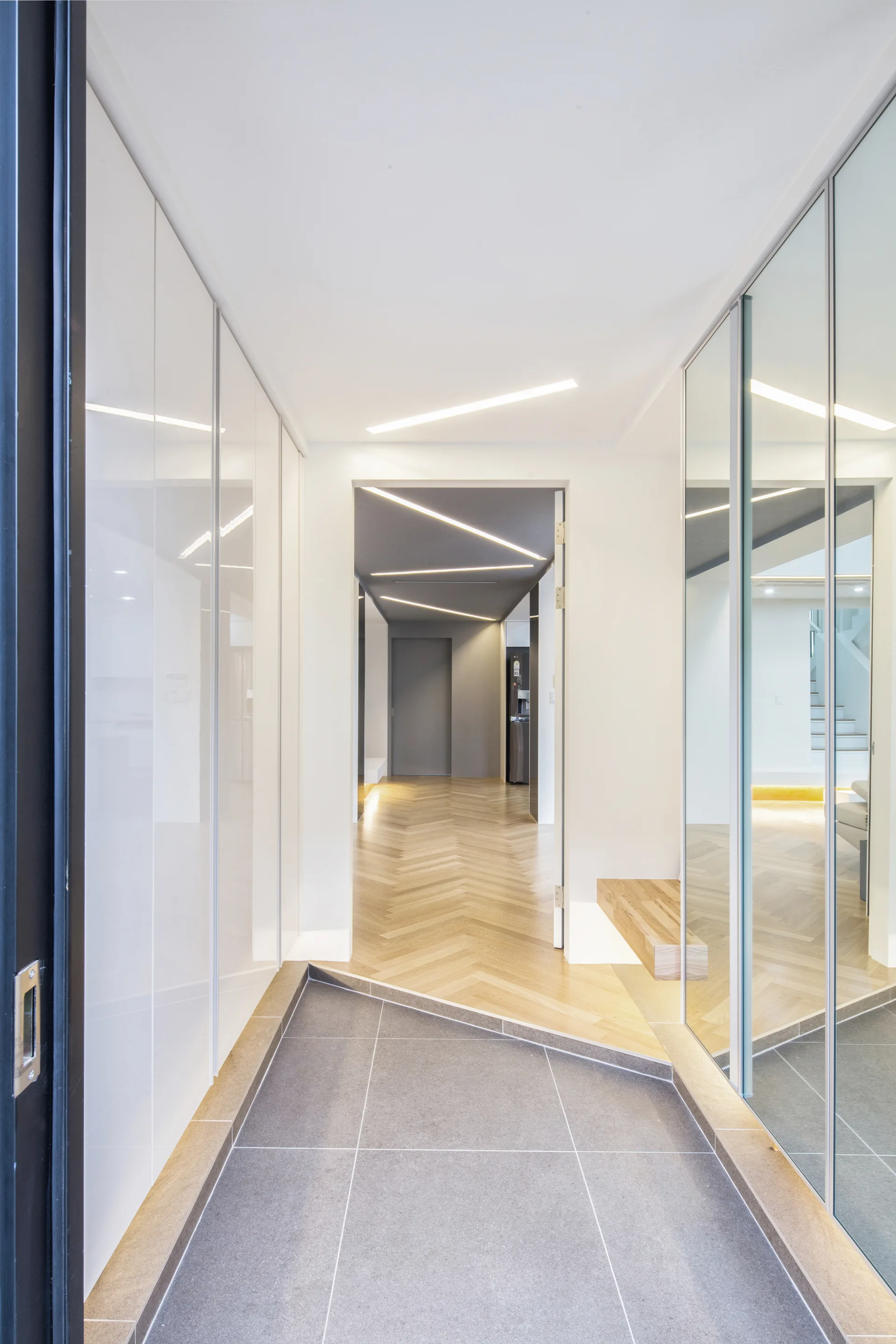 A minimalist modern hallway featuring a diagonal floor transition from gray tile to light wood herringbone flooring alongside mirrored panels.