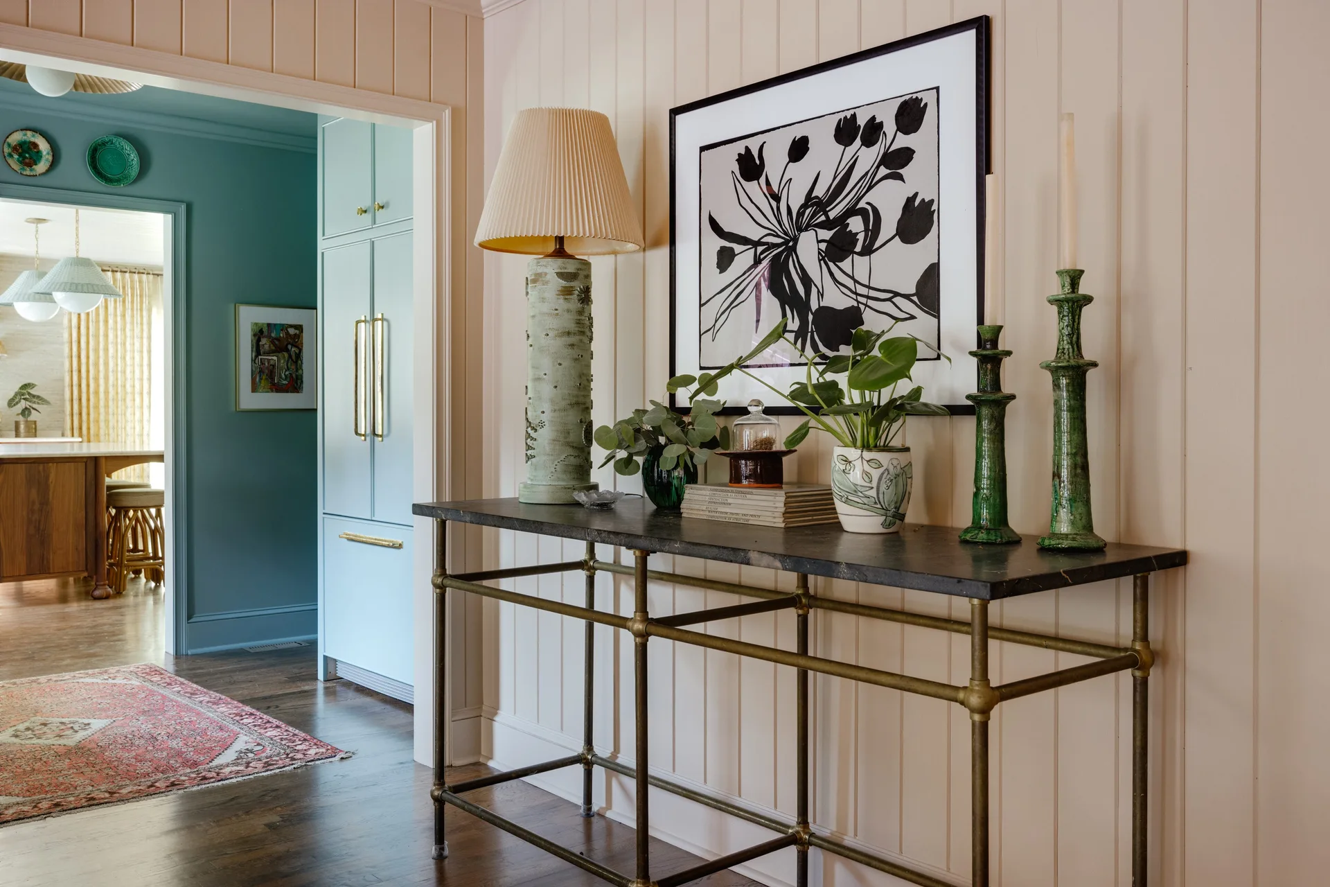 A dark stone console table with brass legs stands against blush-colored vertical wood paneling, decorated with a green ceramic lamp and framed art.