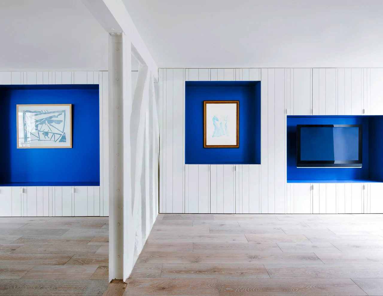 Minimalist room featuring a full wall of white paneled built-in cabinets with three recessed bright blue alcoves holding artwork and a television.