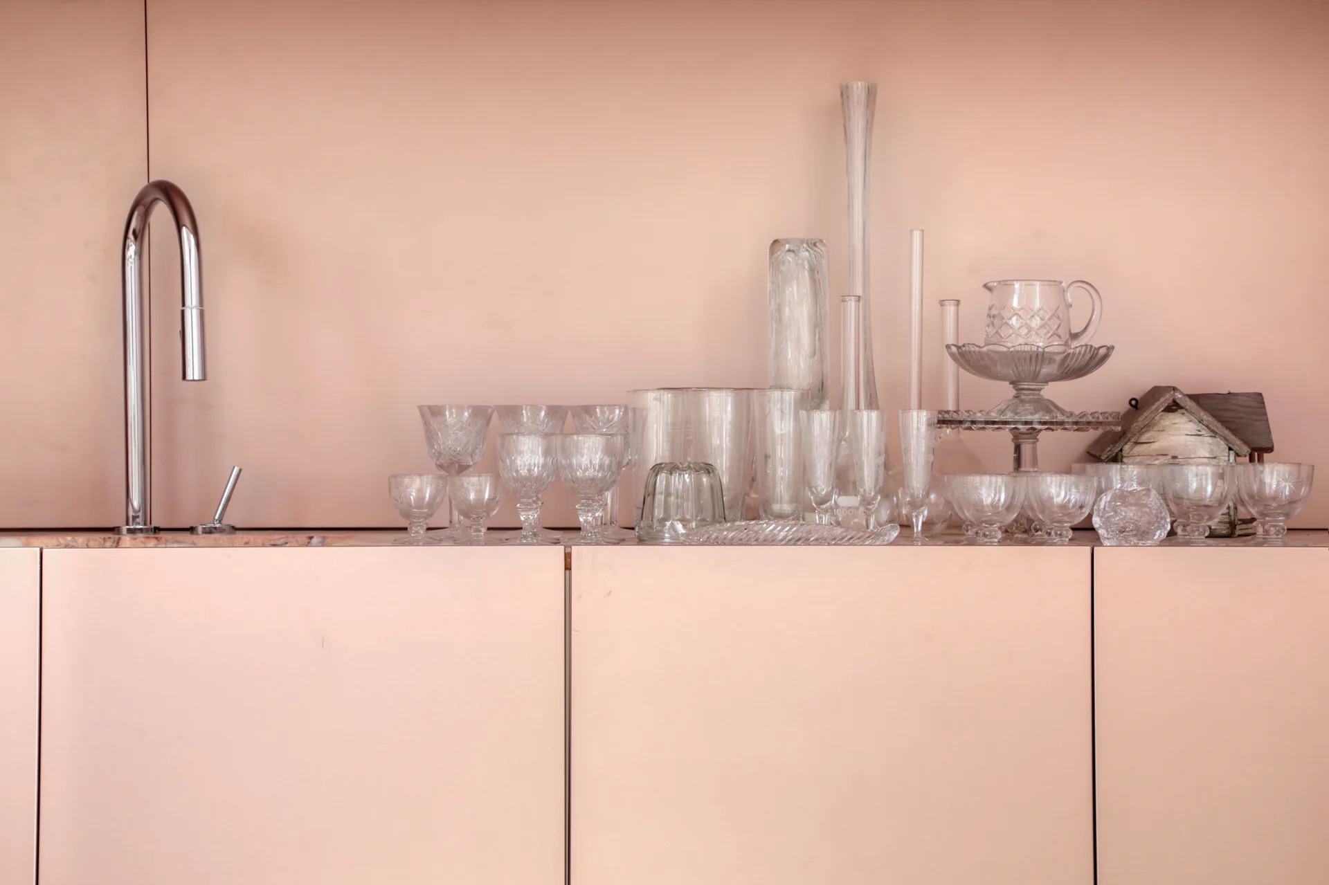 A close-up of matte blush pink kitchen cabinets and matching wall with a chrome faucet and a collection of clear glassware on the countertop.