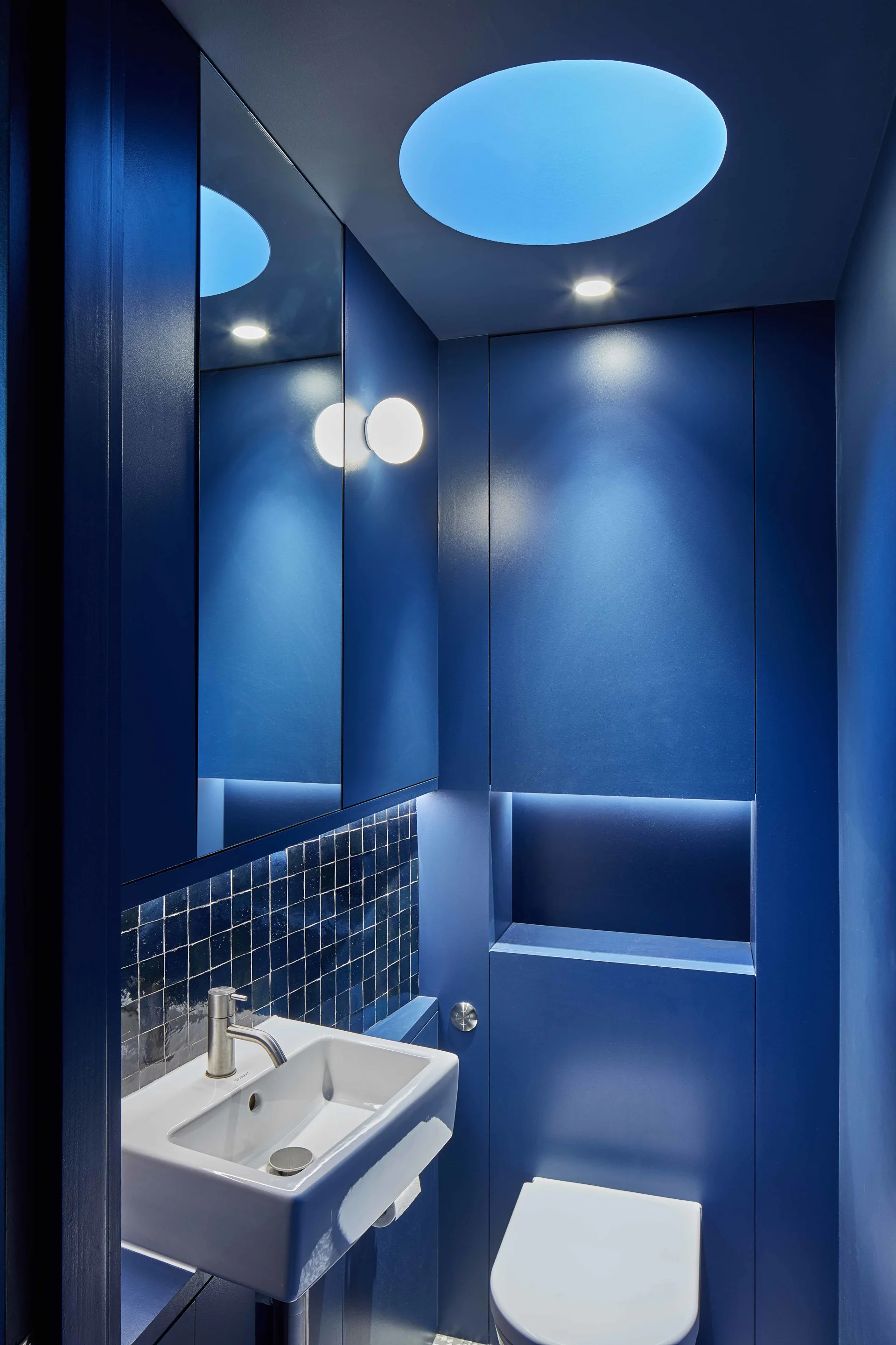 A small navy blue powder room features a white wall-mounted sink, a circular illuminated ceiling element, and seamless built-in wall cabinets.