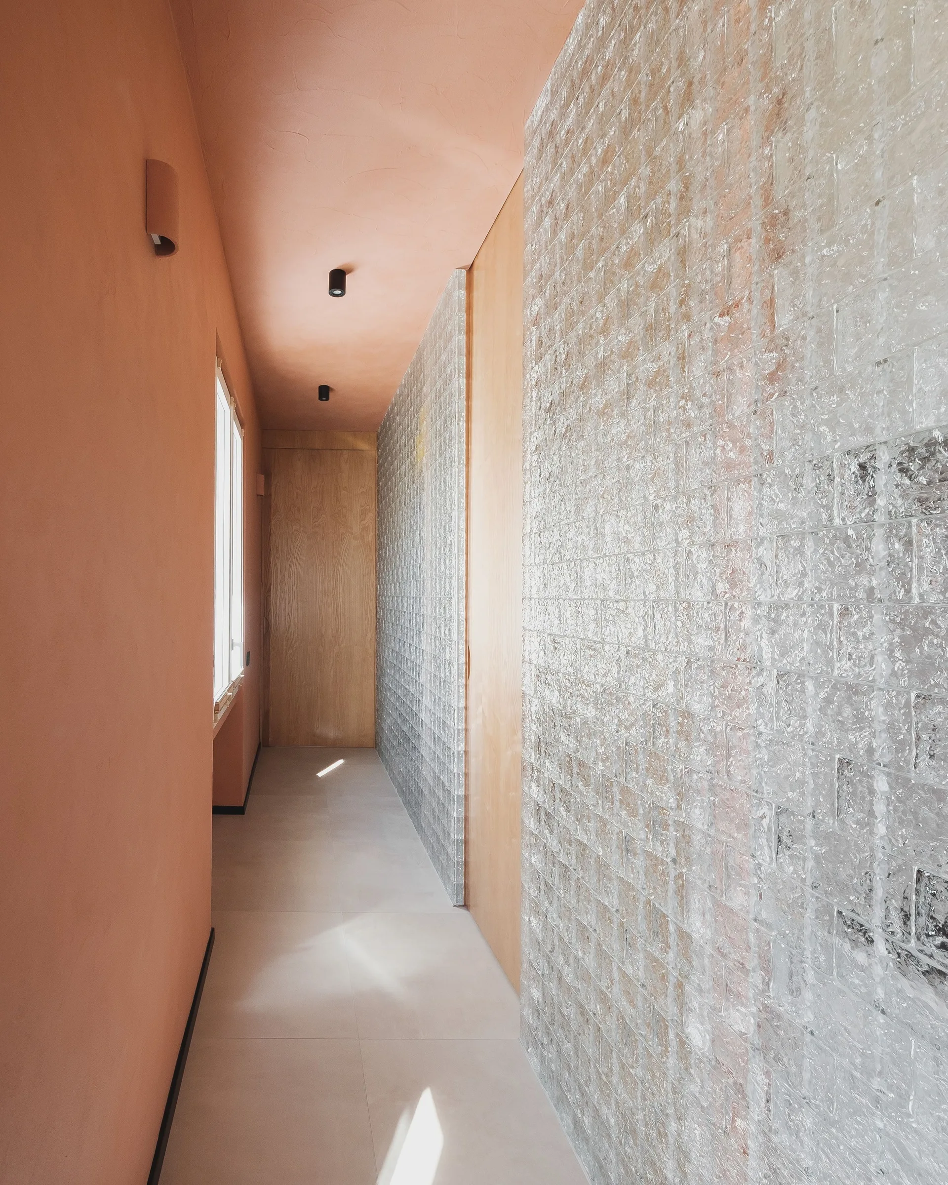 A narrow hallway features a textured glass tile wall, peach-colored plaster ceiling and walls, matte tiled floor, and a light wood door.