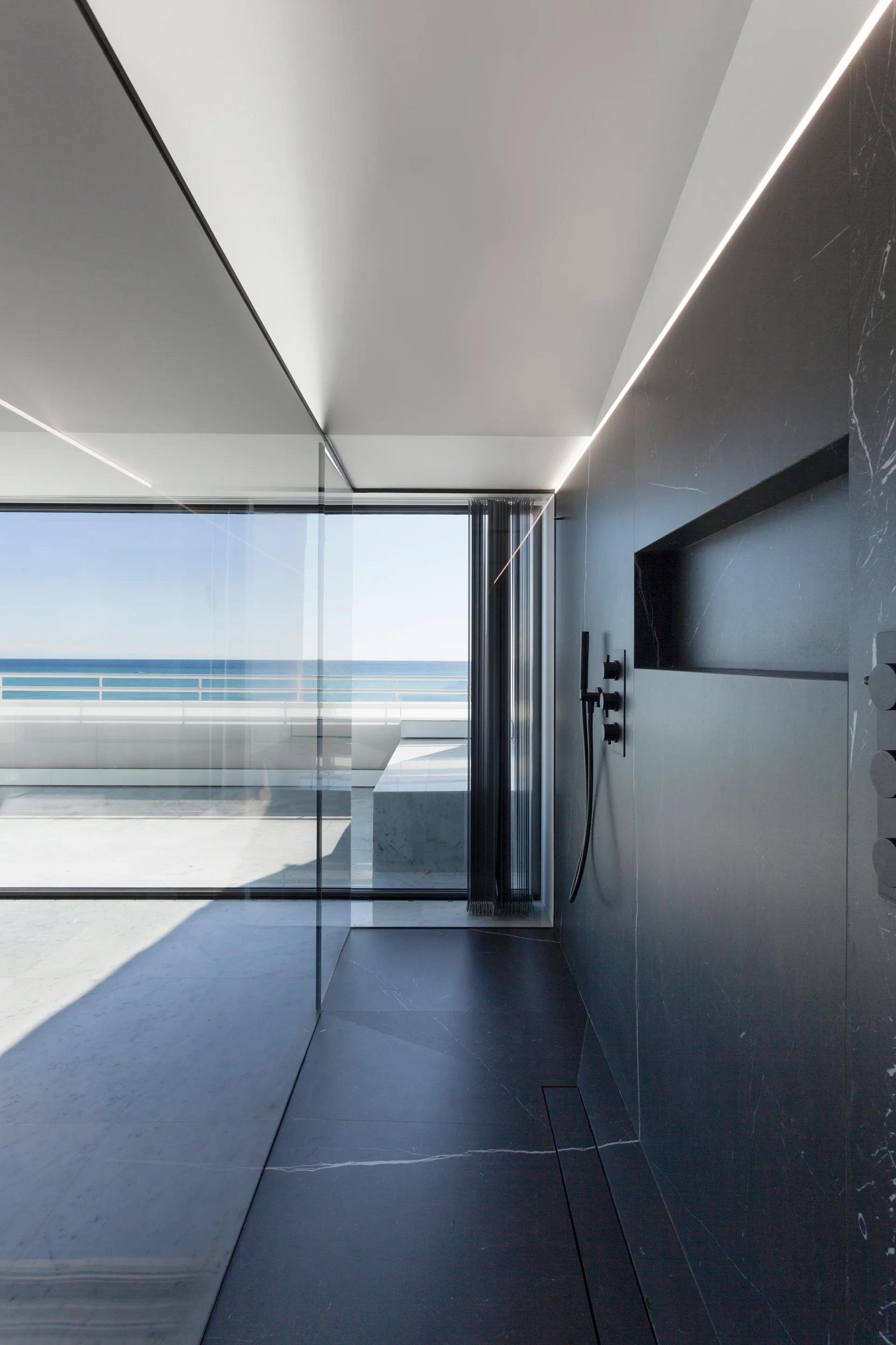 A modern walk-in shower features dark stone-paneled walls, matte black fixtures, a recessed niche, and a glass partition looking out to the ocean.