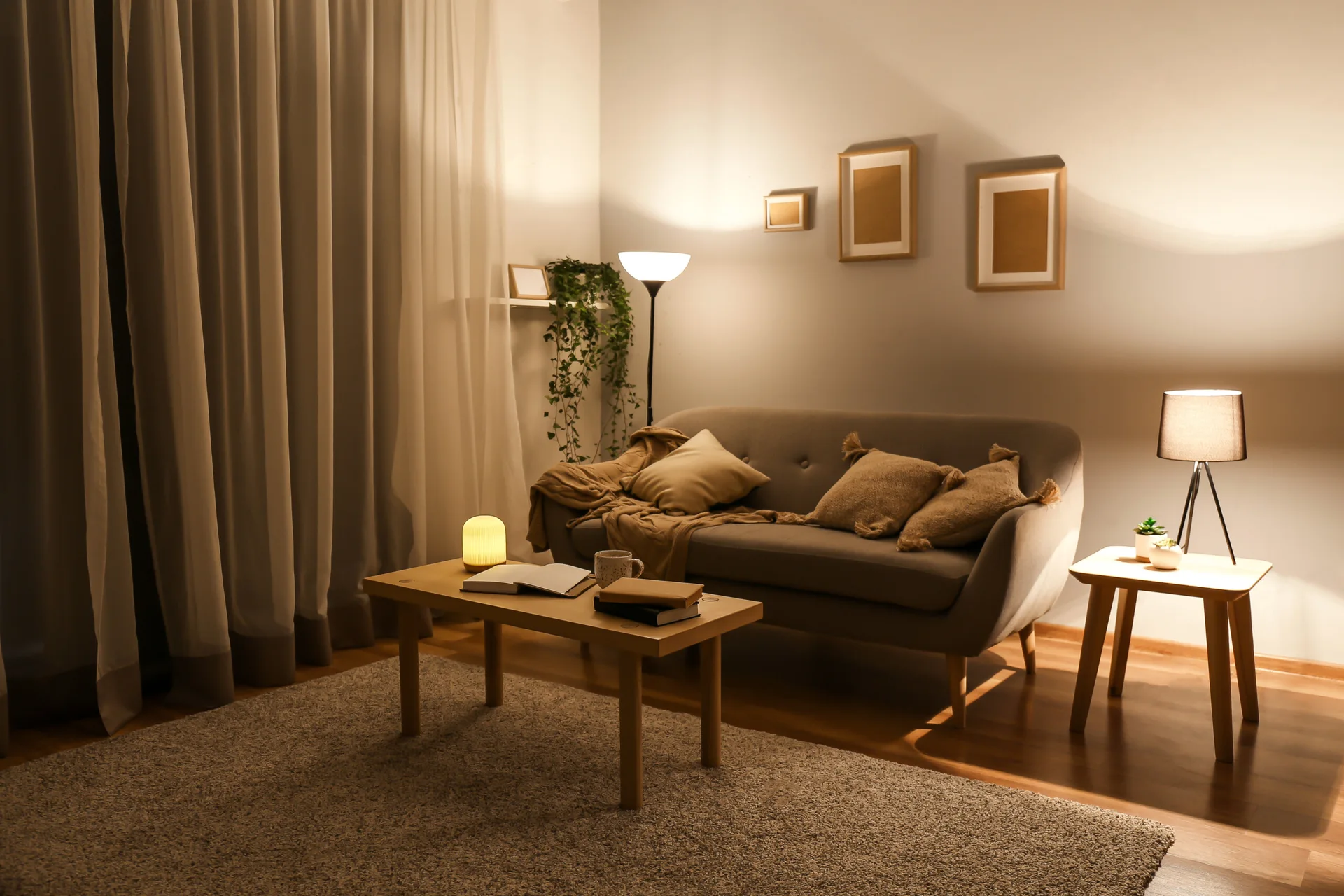 A grey sofa with beige cushions sits in a warmly lit living room next to a wooden coffee table and side table with lamps.