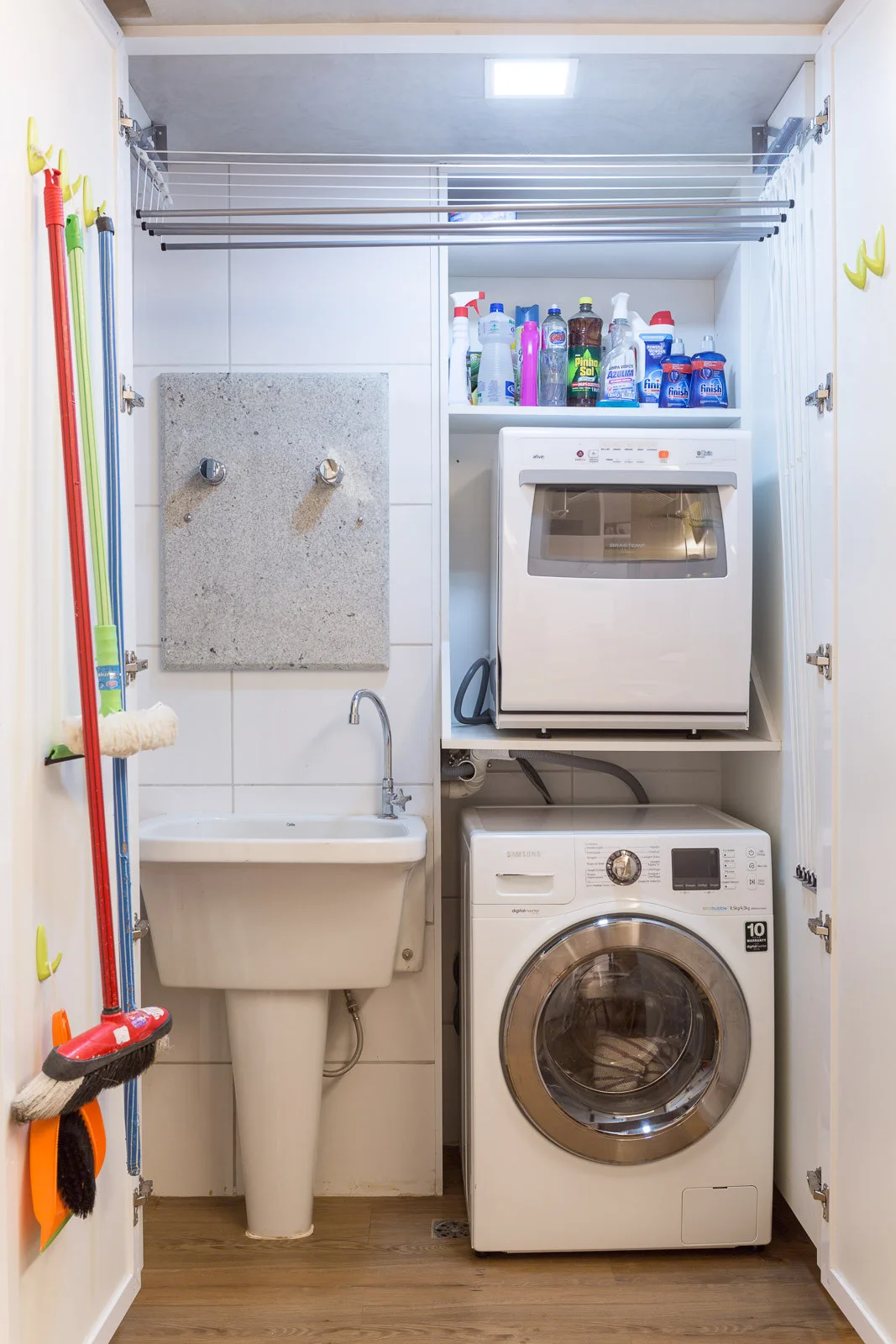 A small laundry closet features a white washing machine, pedestal utility sink, overhead drying rack, and shelves stocked with cleaning supplies.