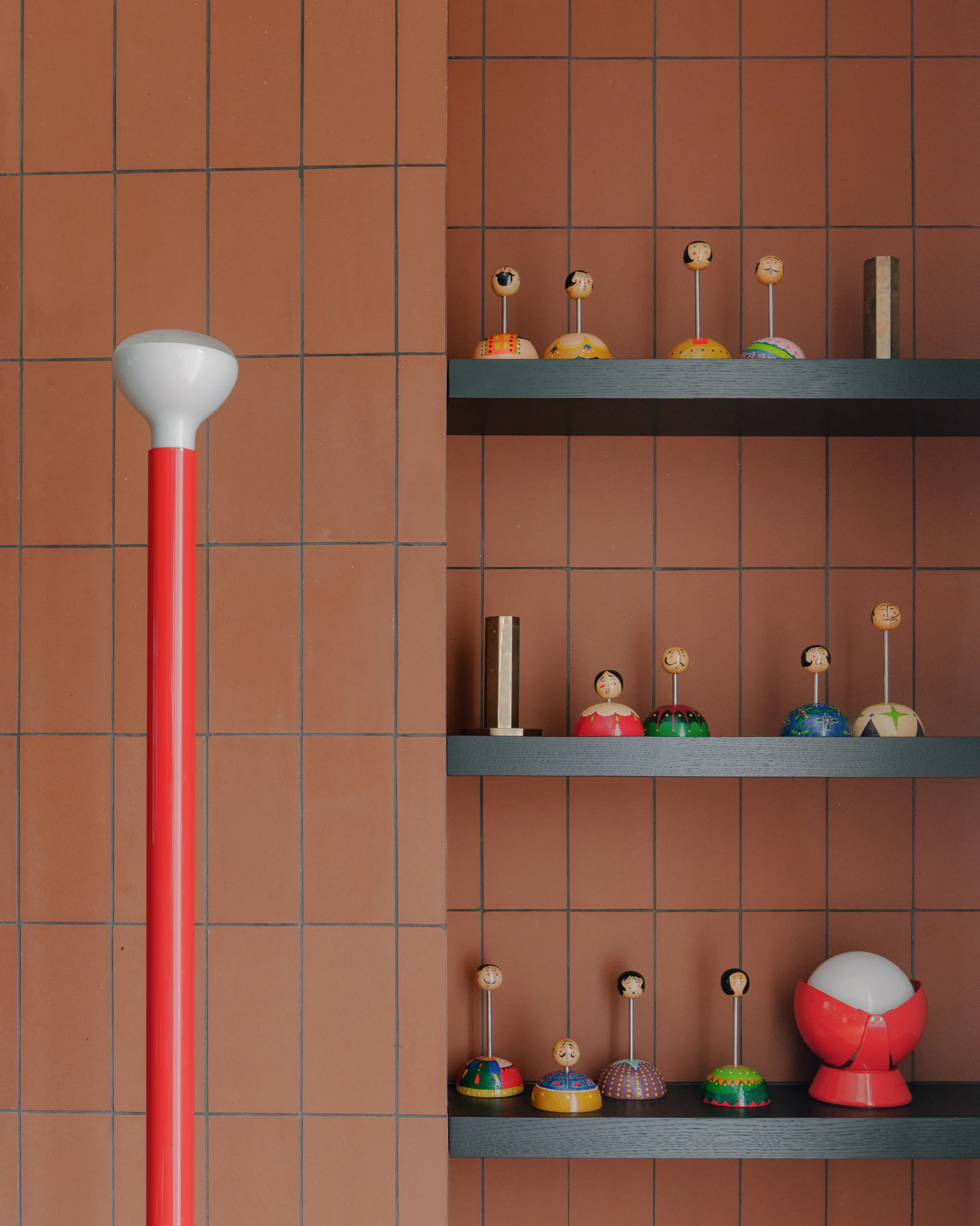 Black recessed shelves display small painted wooden figurines against a terracotta tiled wall next to a tall red lamp.