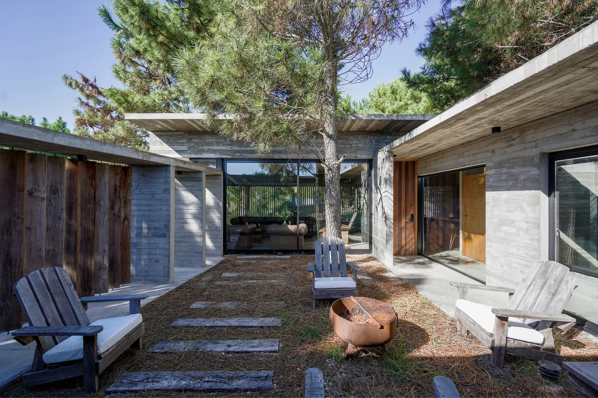 A modern raw concrete courtyard features weathered wooden adirondack chairs, a metal fire pit, and board-formed concrete walls.