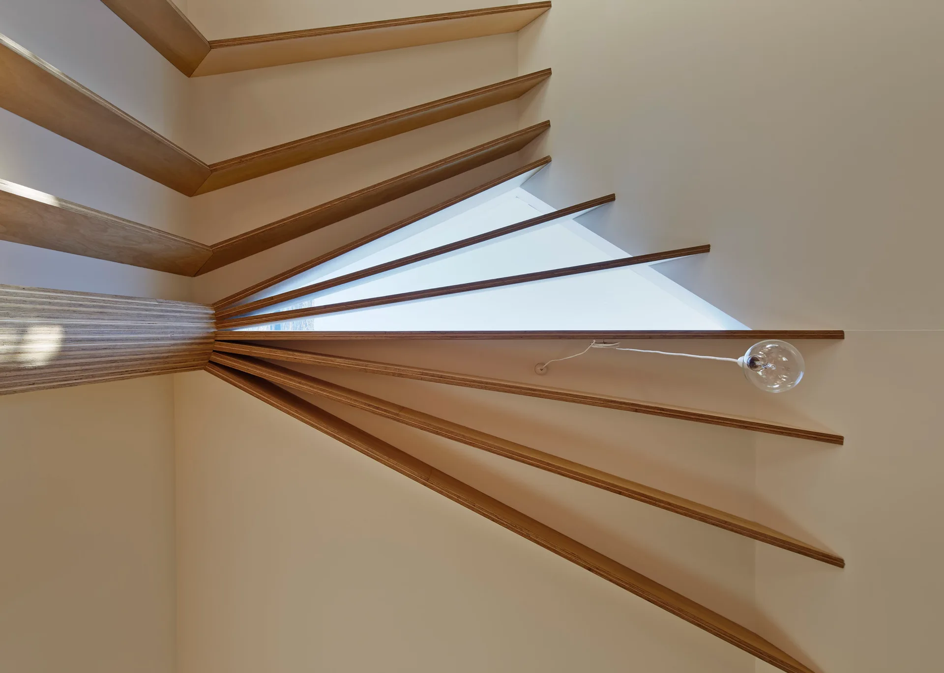 A low-angle shot of a white ceiling featuring radiating light-colored plywood beams converging at a central column next to a triangular skylight.