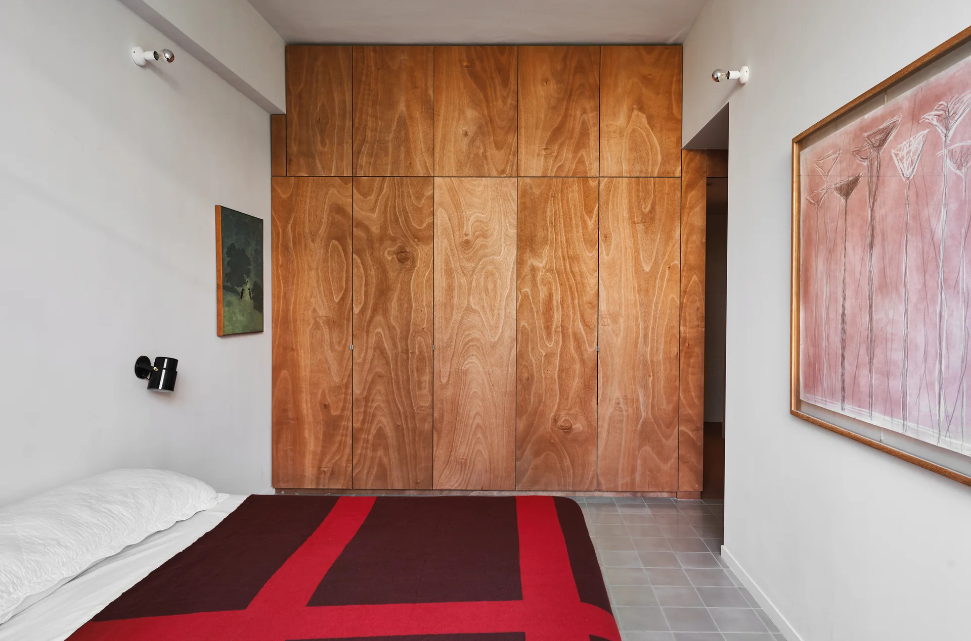 A minimalist bedroom showing a large wood wardrobe wall, a bed with a red and brown blanket, and grey tile flooring.