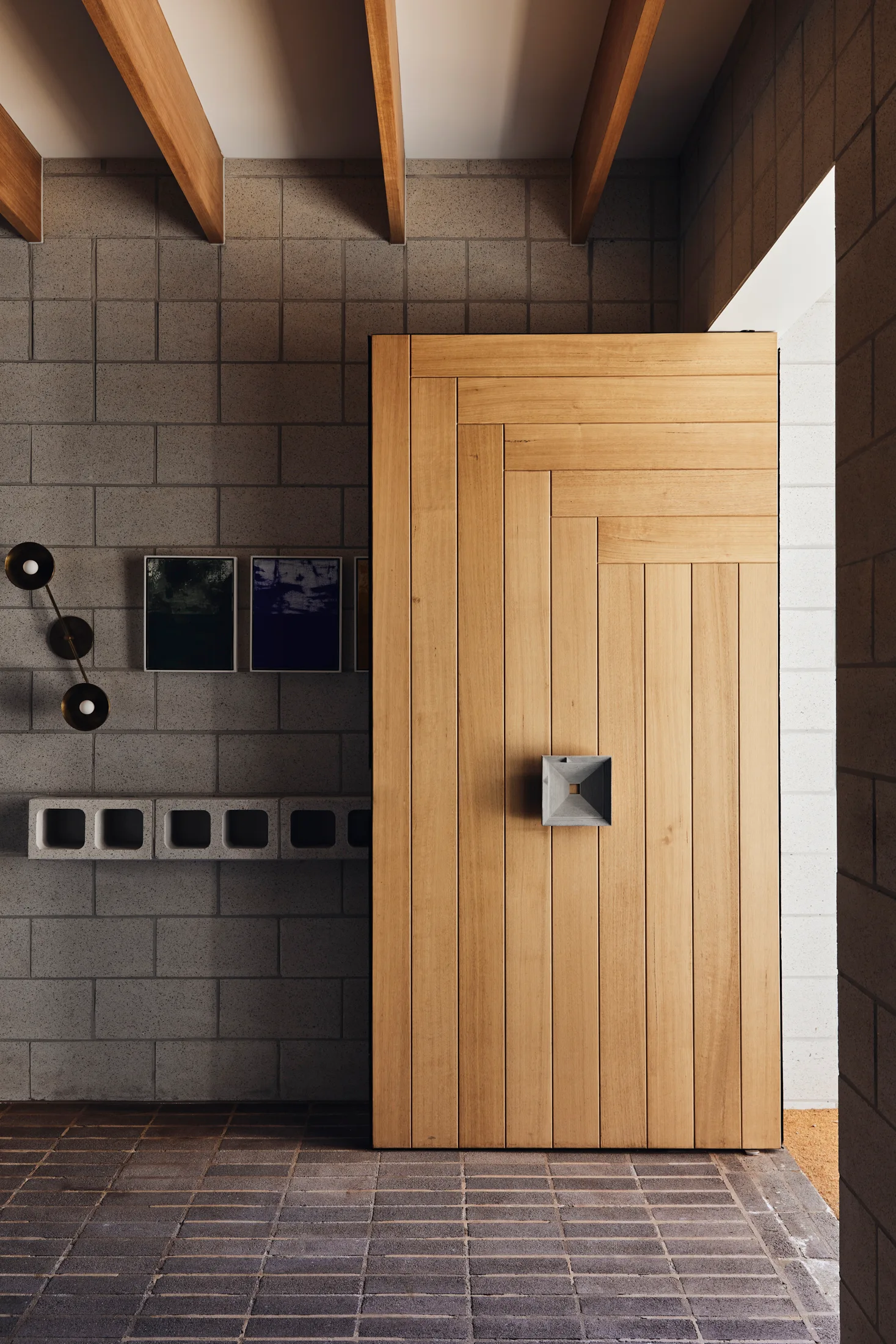 A modern entryway featuring a large geometric wood panel pivot door, grey concrete block walls, and exposed wooden ceiling beams.