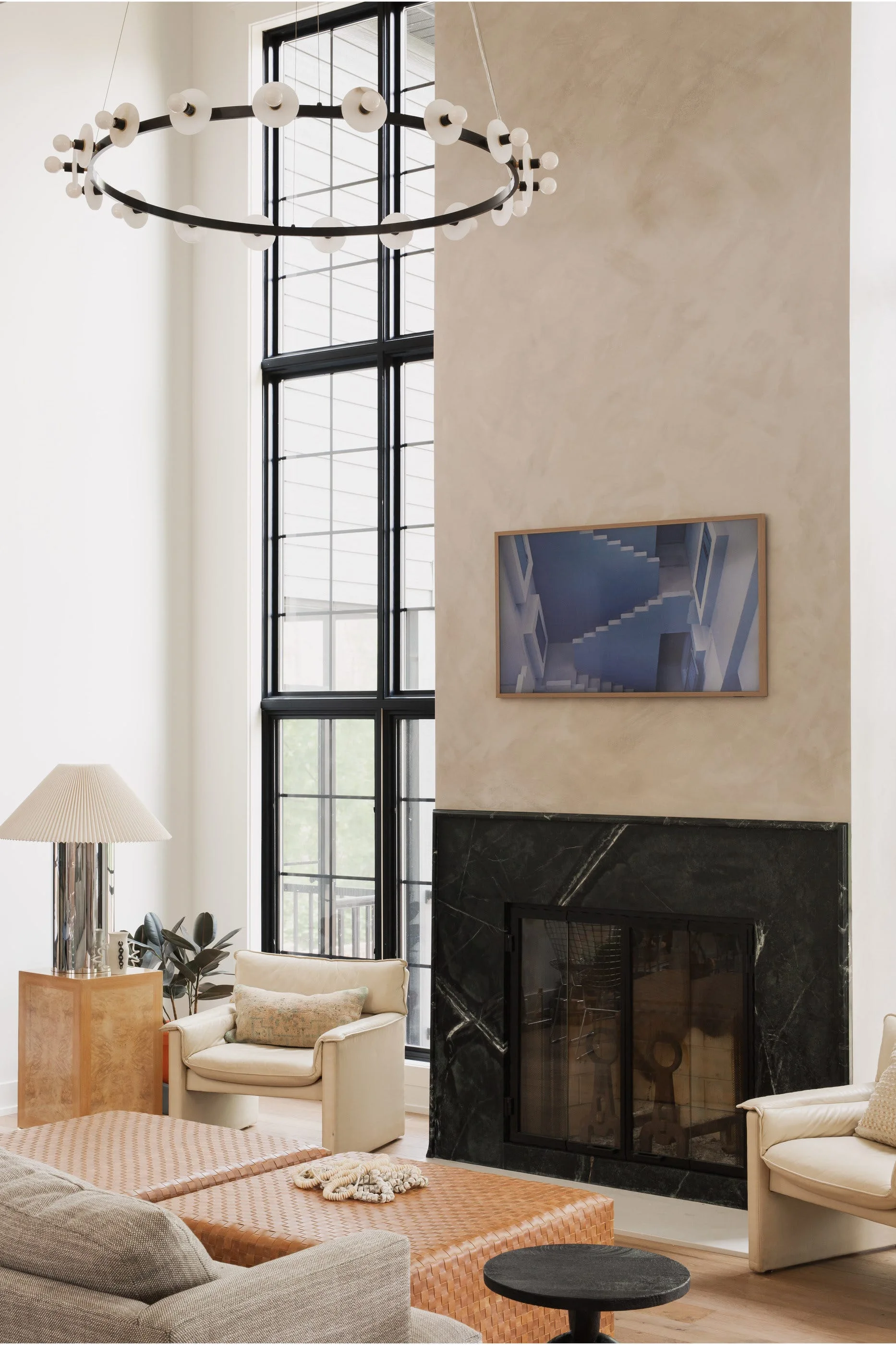 A double-height living room features a dark marble fireplace, cream leather armchairs, a woven leather ottoman, and a large circular ring chandelier.