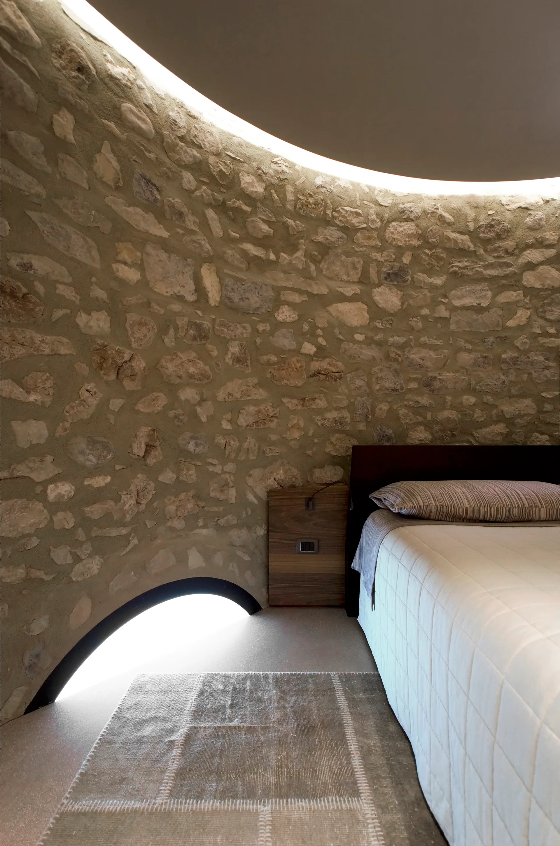 A modern bedroom with a curved natural stone wall, wood nightstand, low-profile bed, and soft cove lighting along the ceiling.