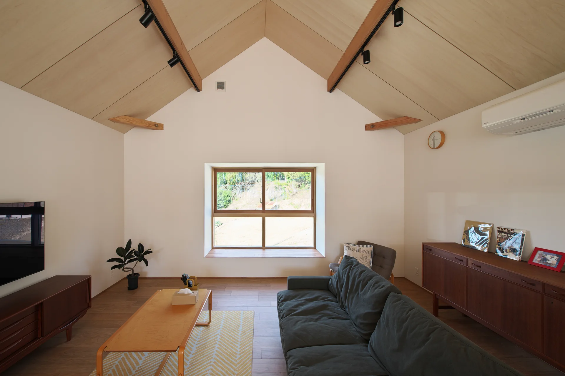 A minimalist living room featuring a vaulted wood-panel ceiling, a dark grey sofa, and a large central window looking out to greenery.