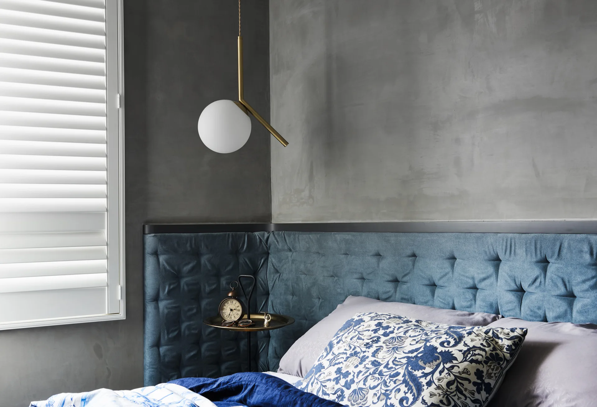 A bedroom corner features a blue tufted headboard against a grey concrete wall, with a gold globe pendant light hanging over a small side table.