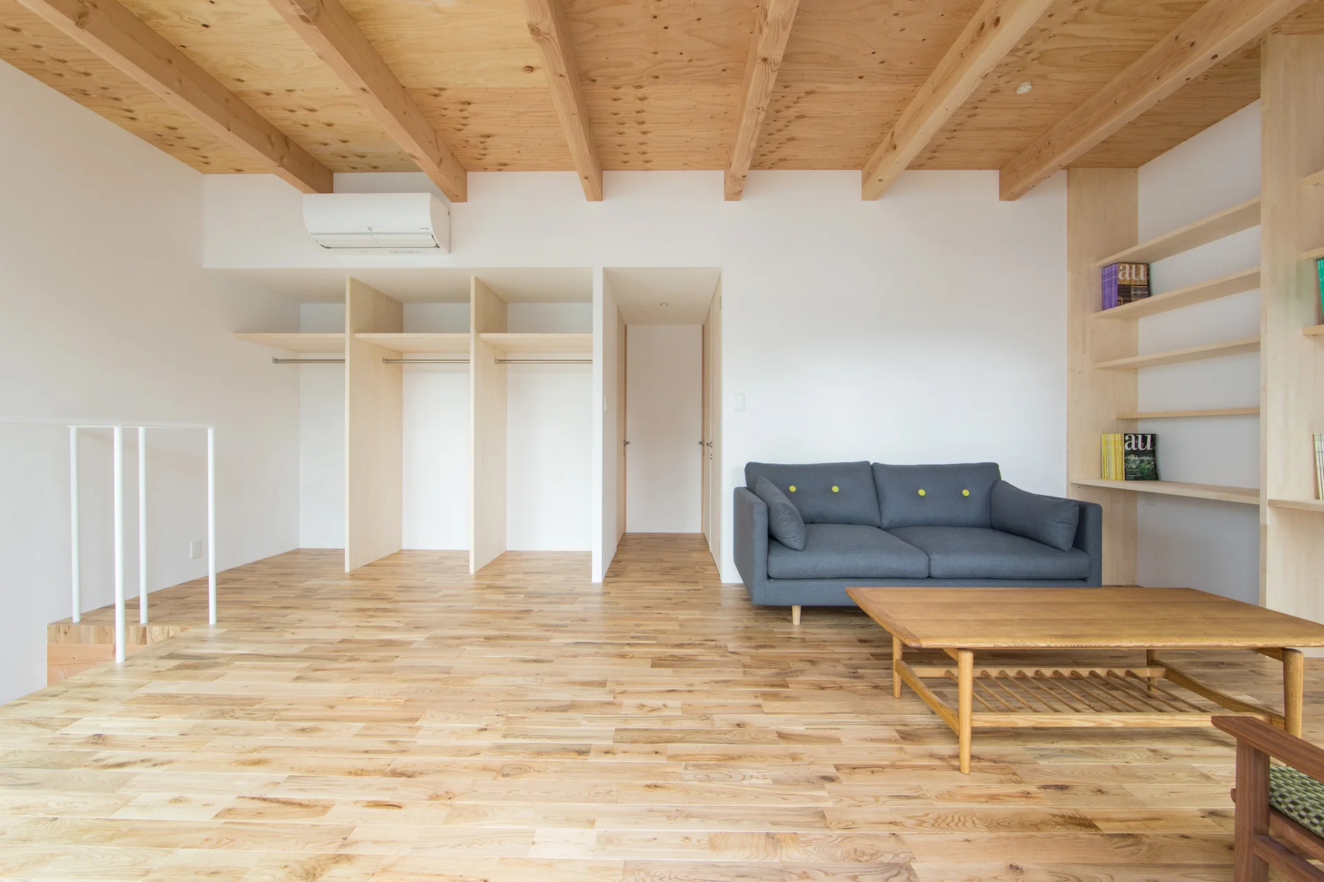 A minimalist living room with light wood floors, exposed ceiling beams, a grey sofa, and open built-in shelving.