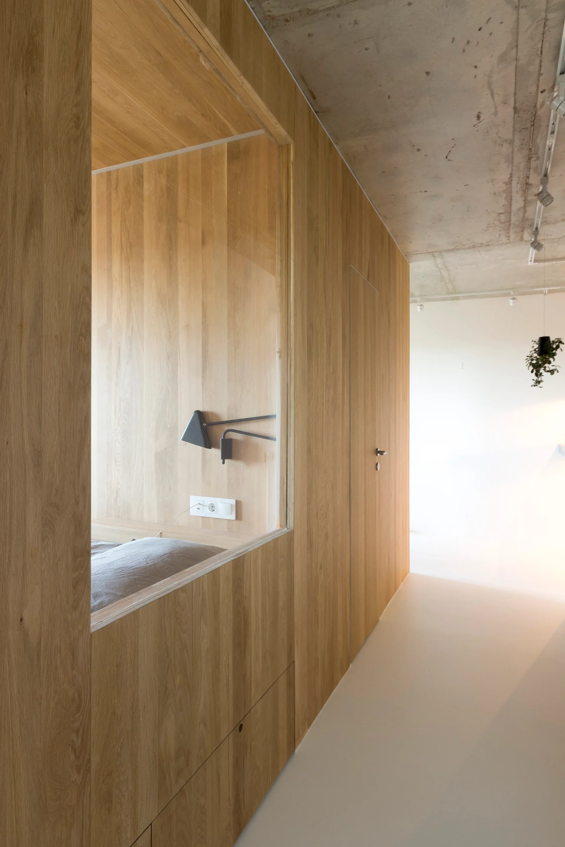 A minimalist hallway features a light oak partition wall with an integrated glass window revealing a built-in bed nook.