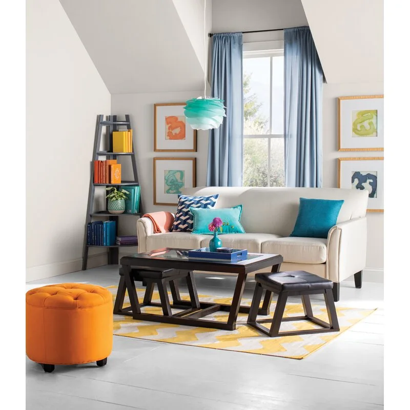 A bright living room with a beige sofa, blue curtains, a dark wood coffee table with nesting stools, and a tufted orange ottoman.