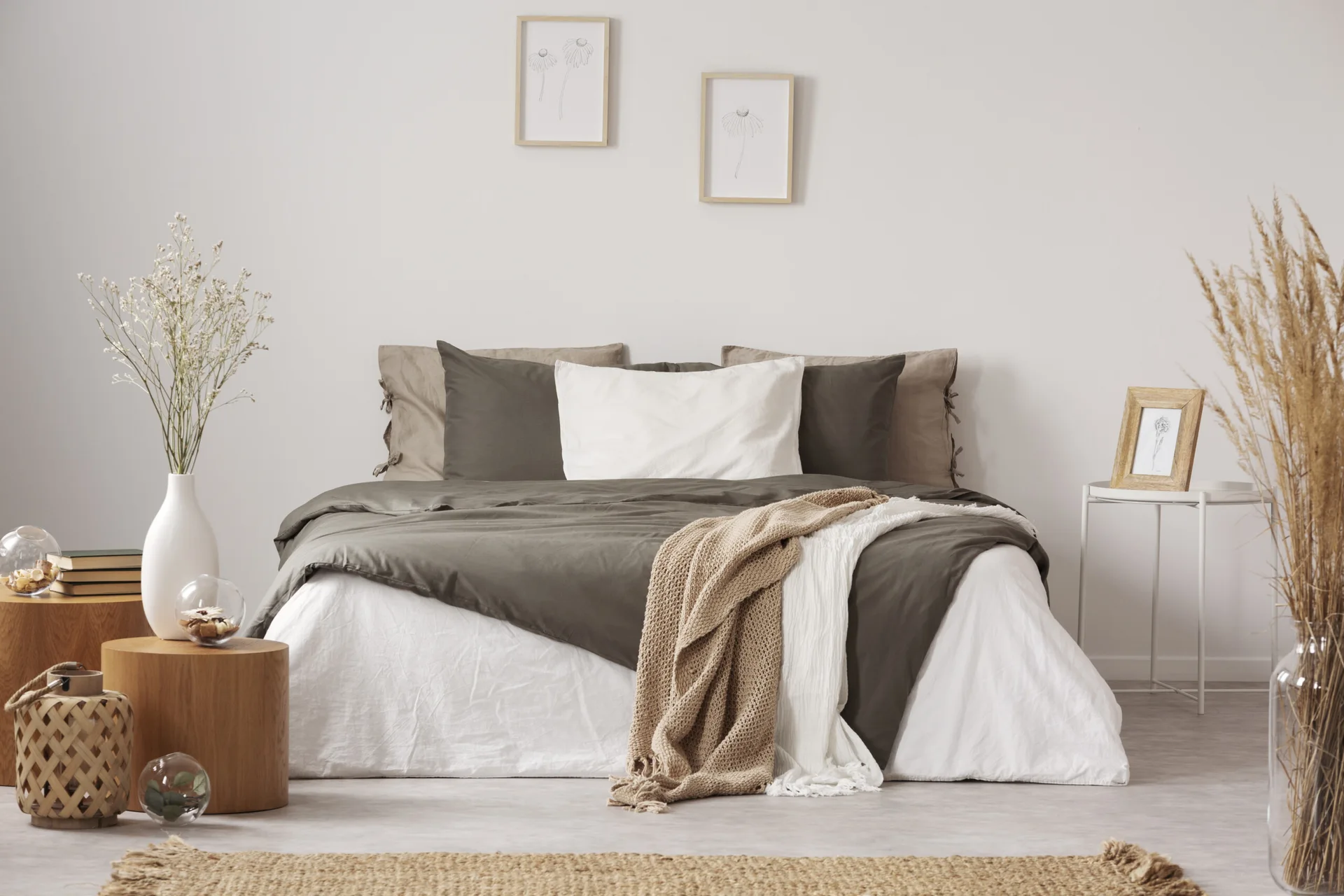 A minimalist bedroom features a bed with olive green and white linens, flanked by cylindrical wooden side tables and a light-colored wall with framed art.