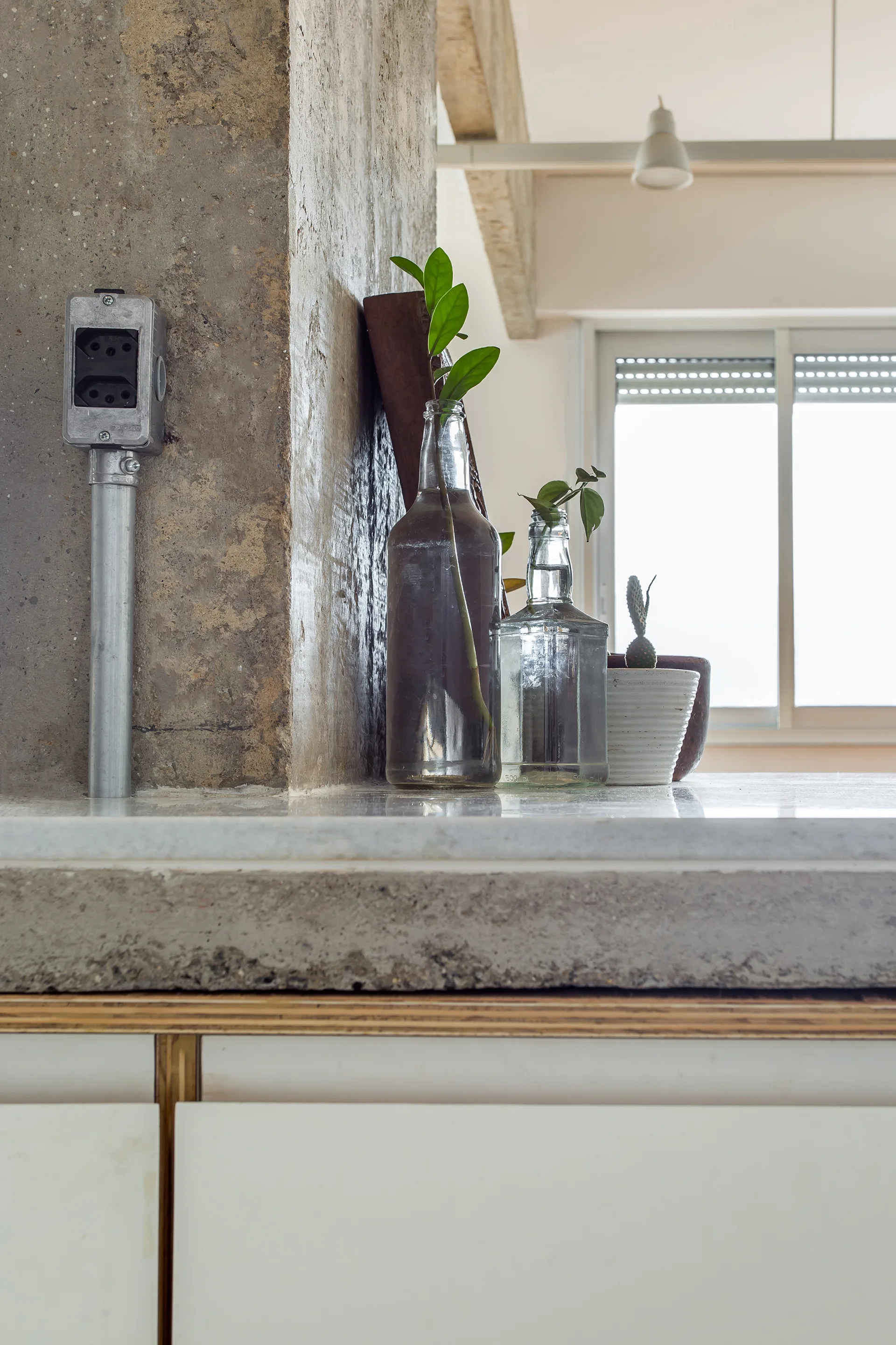 A close-up of a concrete kitchen countertop with glass bottle vases, a small potted cactus, and a raw concrete pillar with metal conduit.