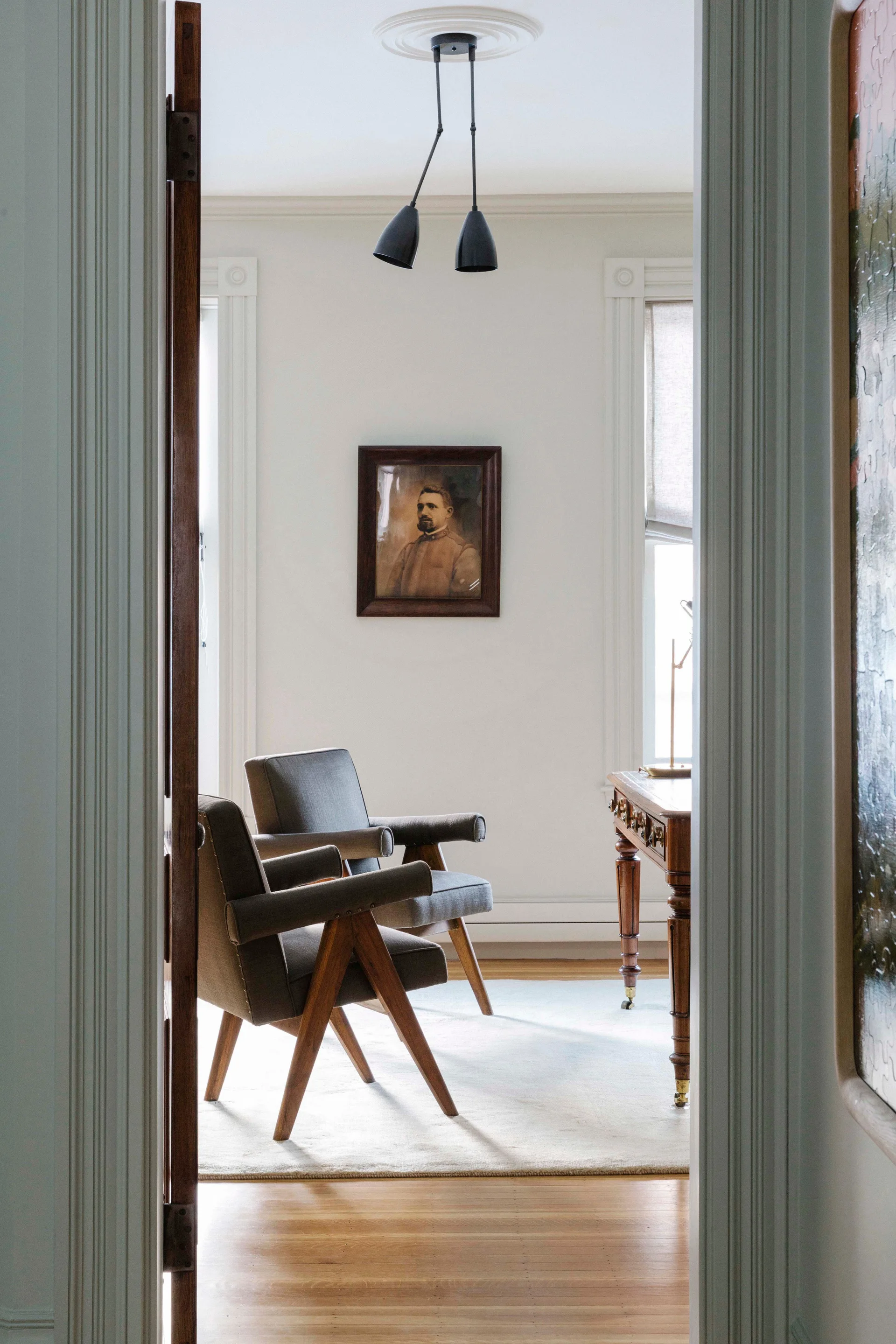 A view through a doorway into a home office featuring two mid-century armchairs, a wooden desk, and a vintage portrait.