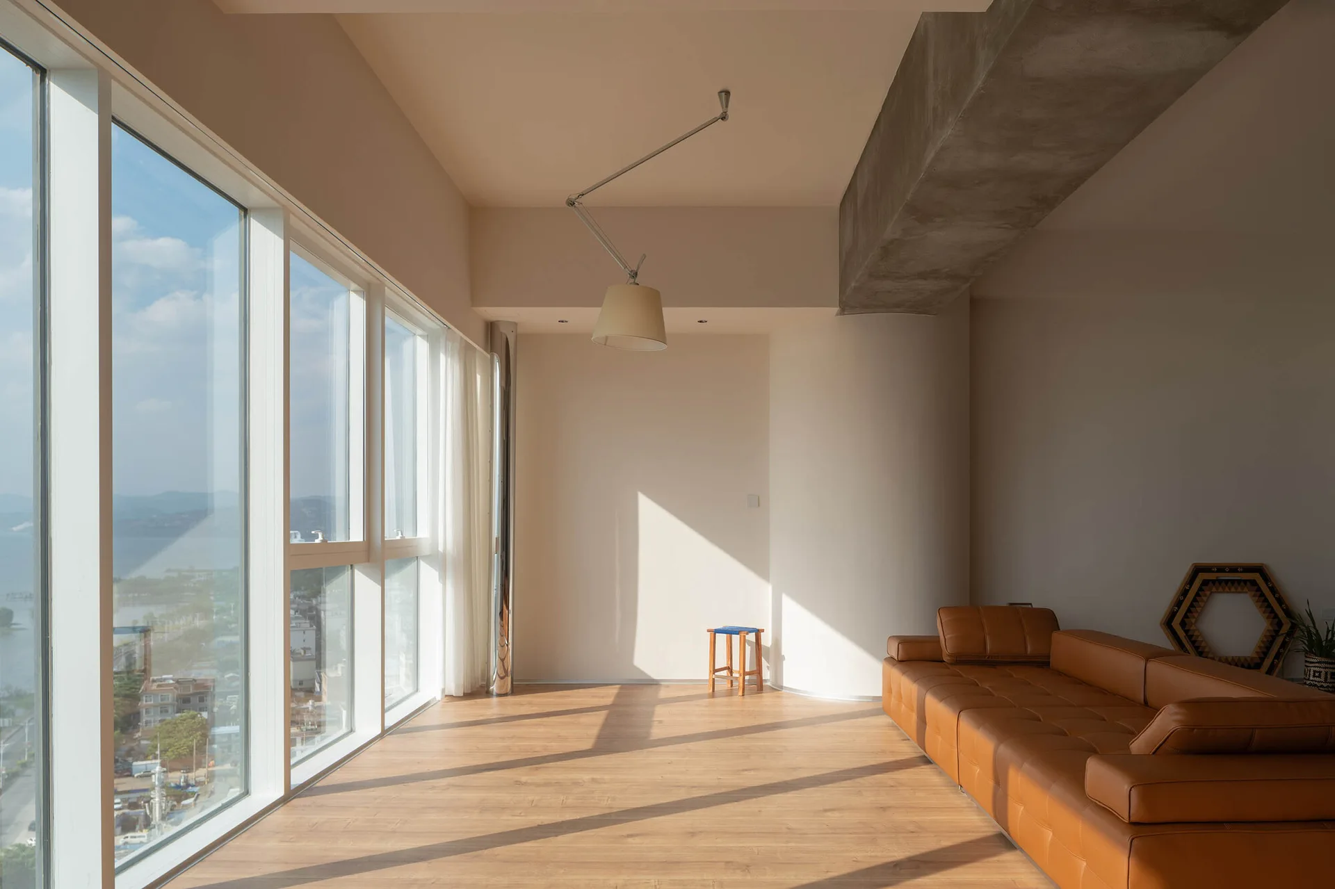 A minimalist living room features a tan leather sofa, an exposed concrete ceiling beam, and floor-to-ceiling windows showing a bright outdoor landscape.