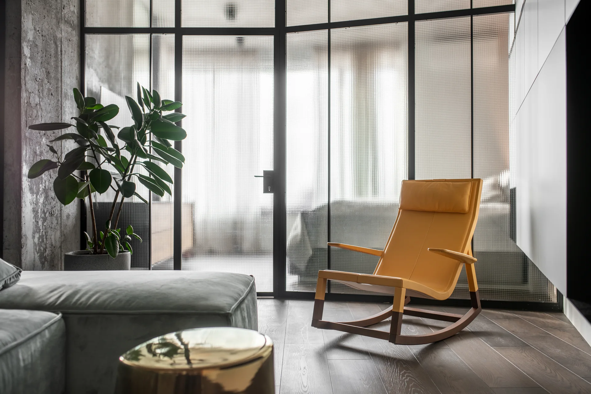 A modern living space features a yellow leather rocking chair and a grey sofa next to a black-framed reeded glass room partition.