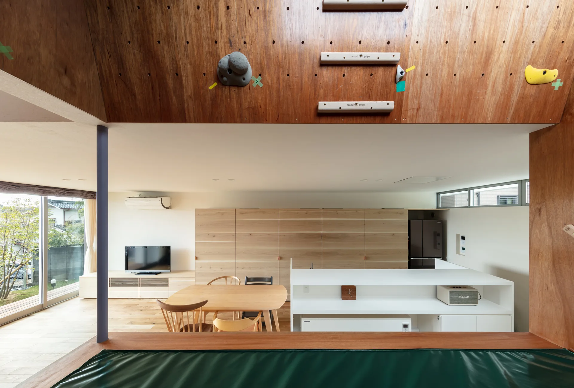 A modern open-plan kitchen and dining area with a wooden table, light wood cabinetry, and an overhead wooden structure fitted with climbing holds.