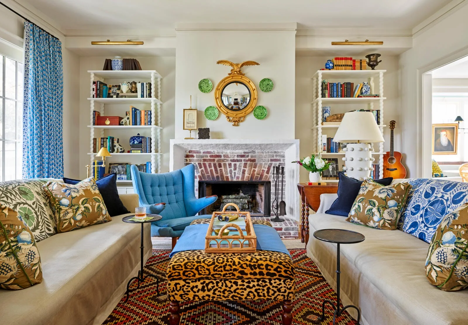 An eclectic living room featuring facing beige sofas, a leopard-print ottoman, a brick fireplace, and white bookshelves.