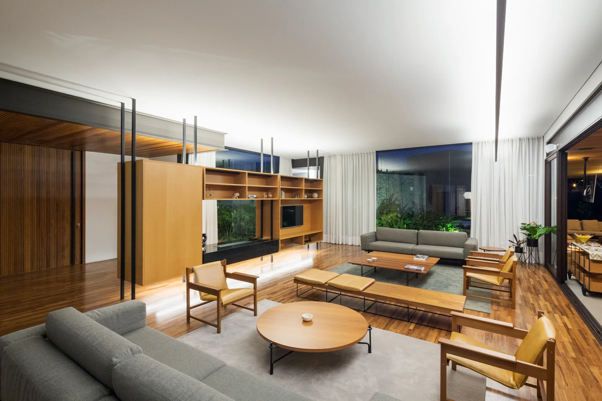A spacious living room featuring gray fabric sofas, wooden armchairs, a large wood shelving divider, and floor-to-ceiling windows.