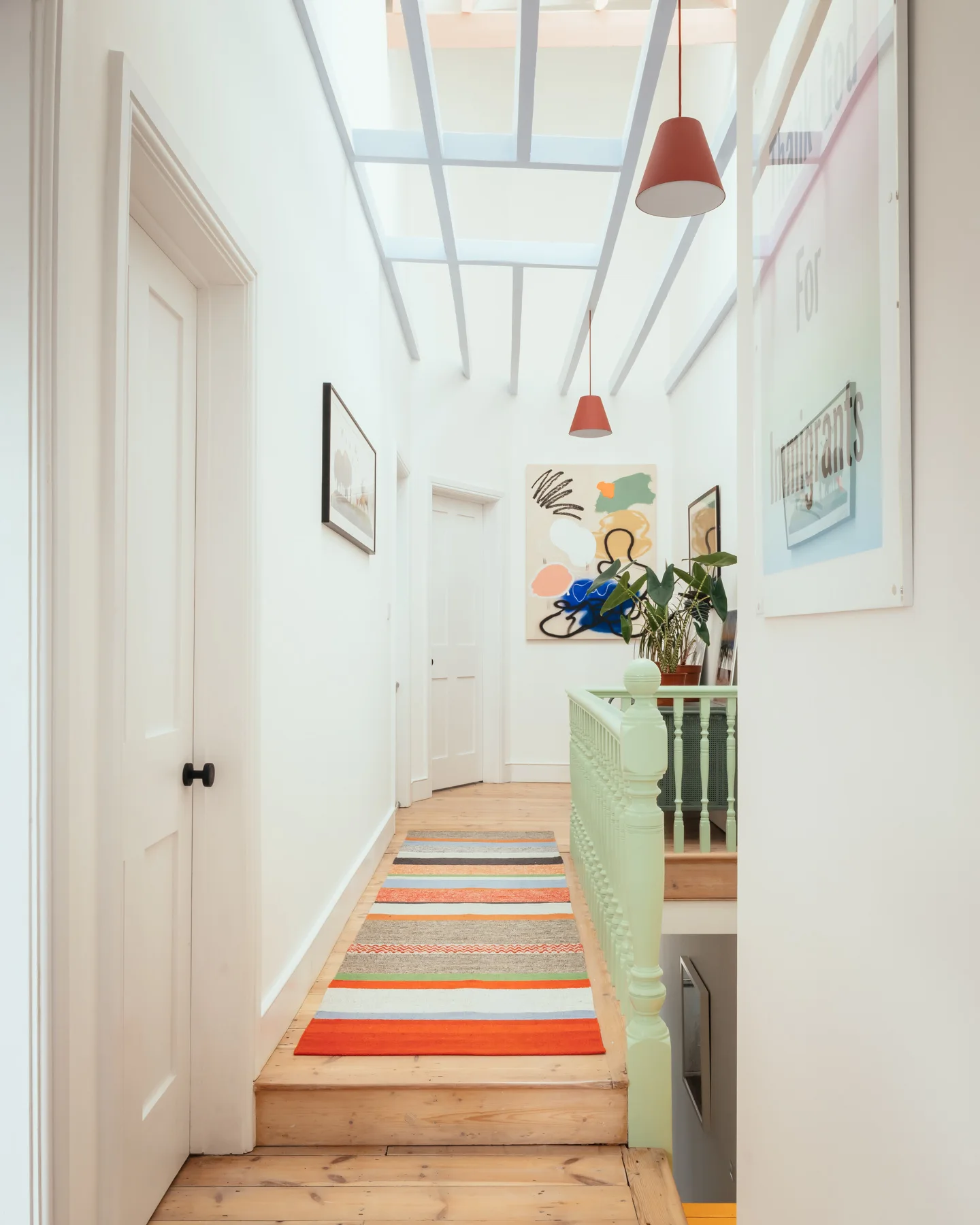 A bright white hallway features a colorful striped runner rug on natural wood floors, a light green balustrade, and a skylight ceiling grid.