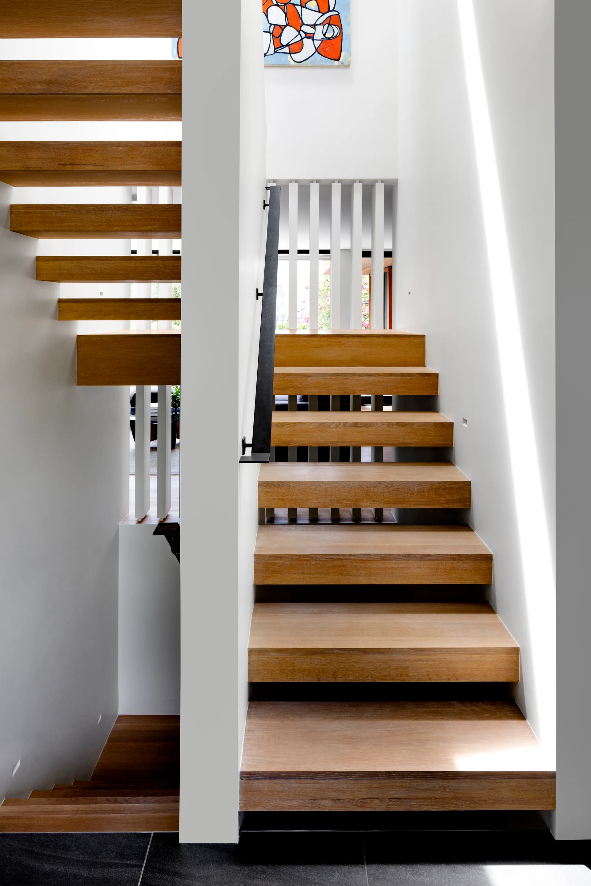 A modern split staircase features thick natural wood treads, white walls, vertical white balusters, and a black metal handrail.