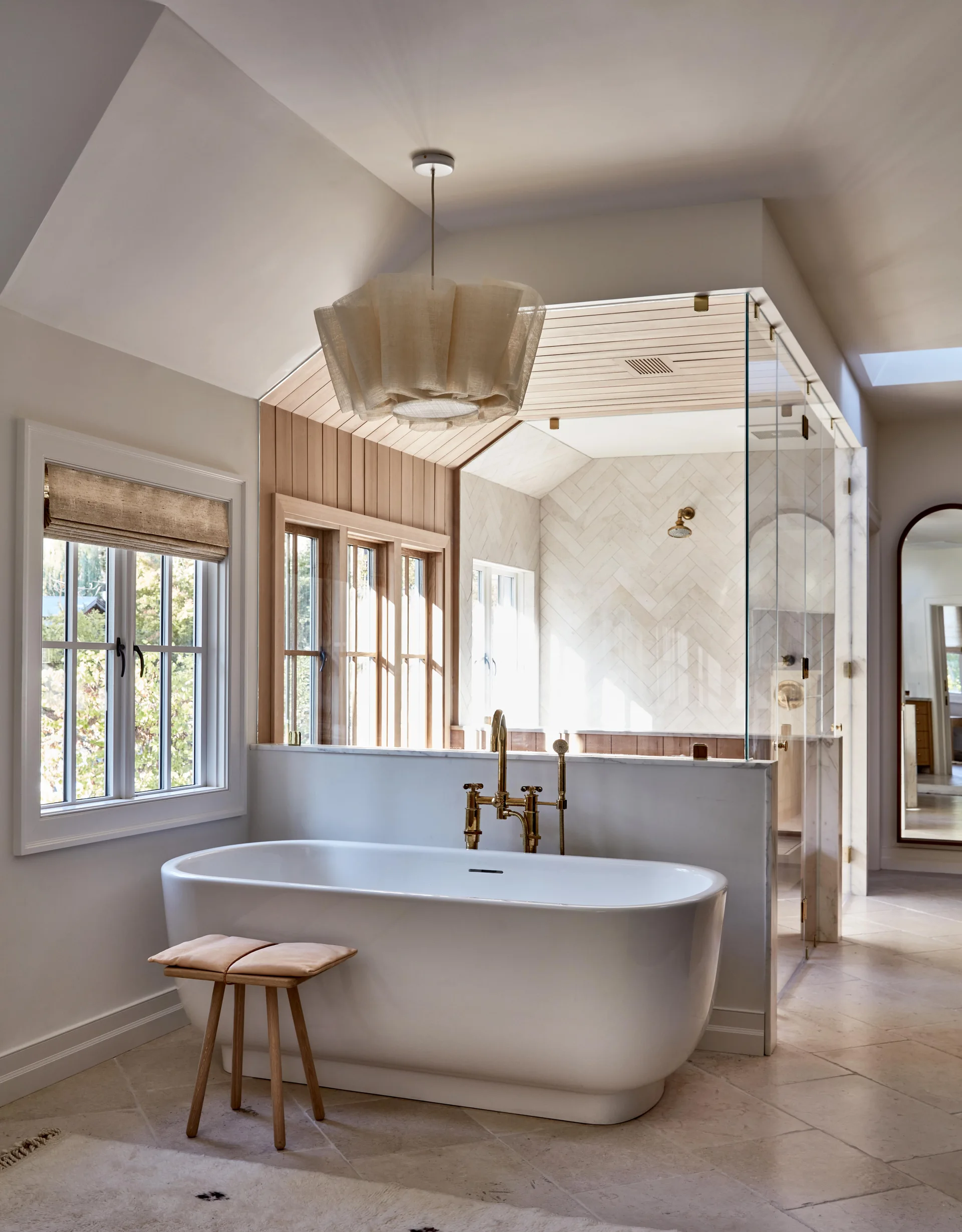 A bright bathroom features a white freestanding bathtub with brass fixtures next to a glass-enclosed shower with beige herringbone tiling.