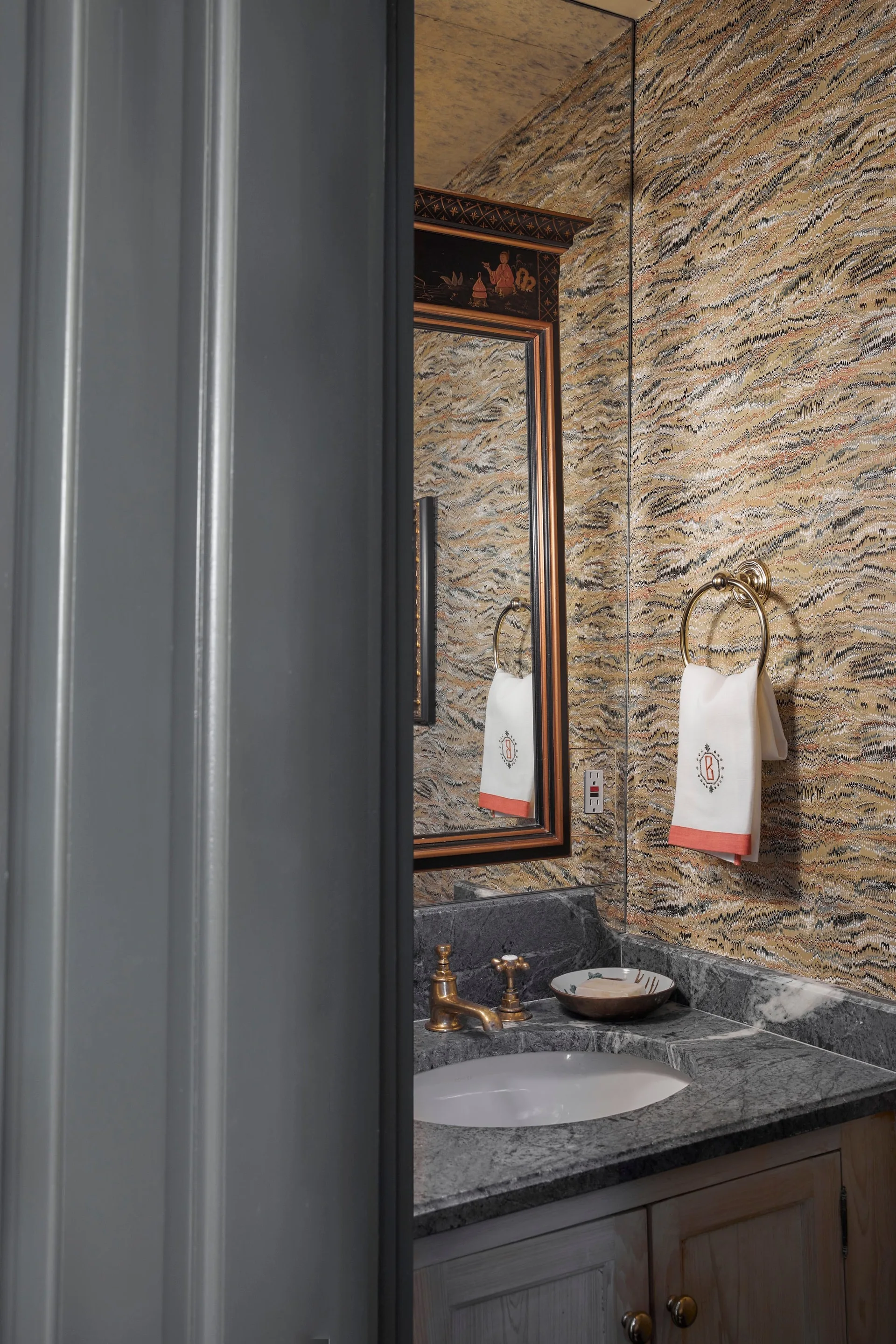 A powder room features a gray marble vanity with a brass faucet, a monogrammed hand towel on a brass ring, and patterned marbled wallpaper.