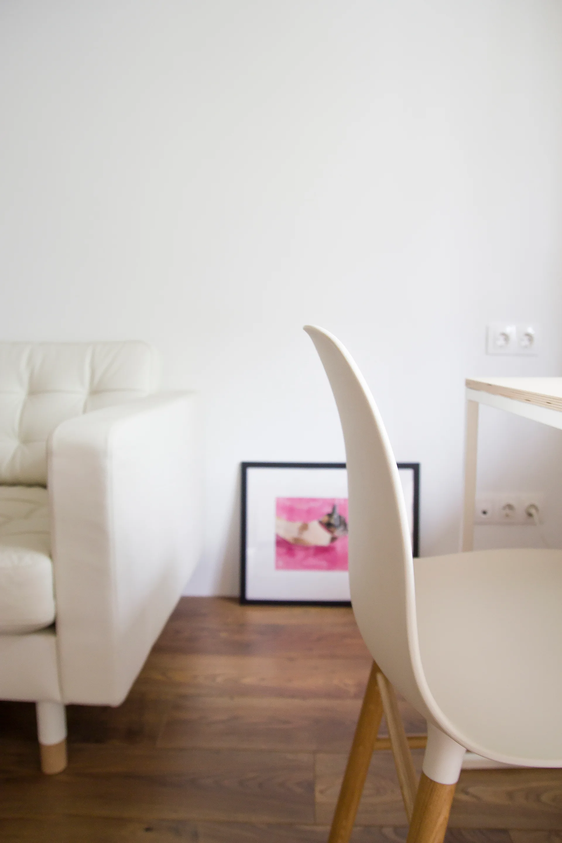 A white tufted sofa and a modern white chair sit on a dark wood floor near a small pink framed artwork resting against a white wall.