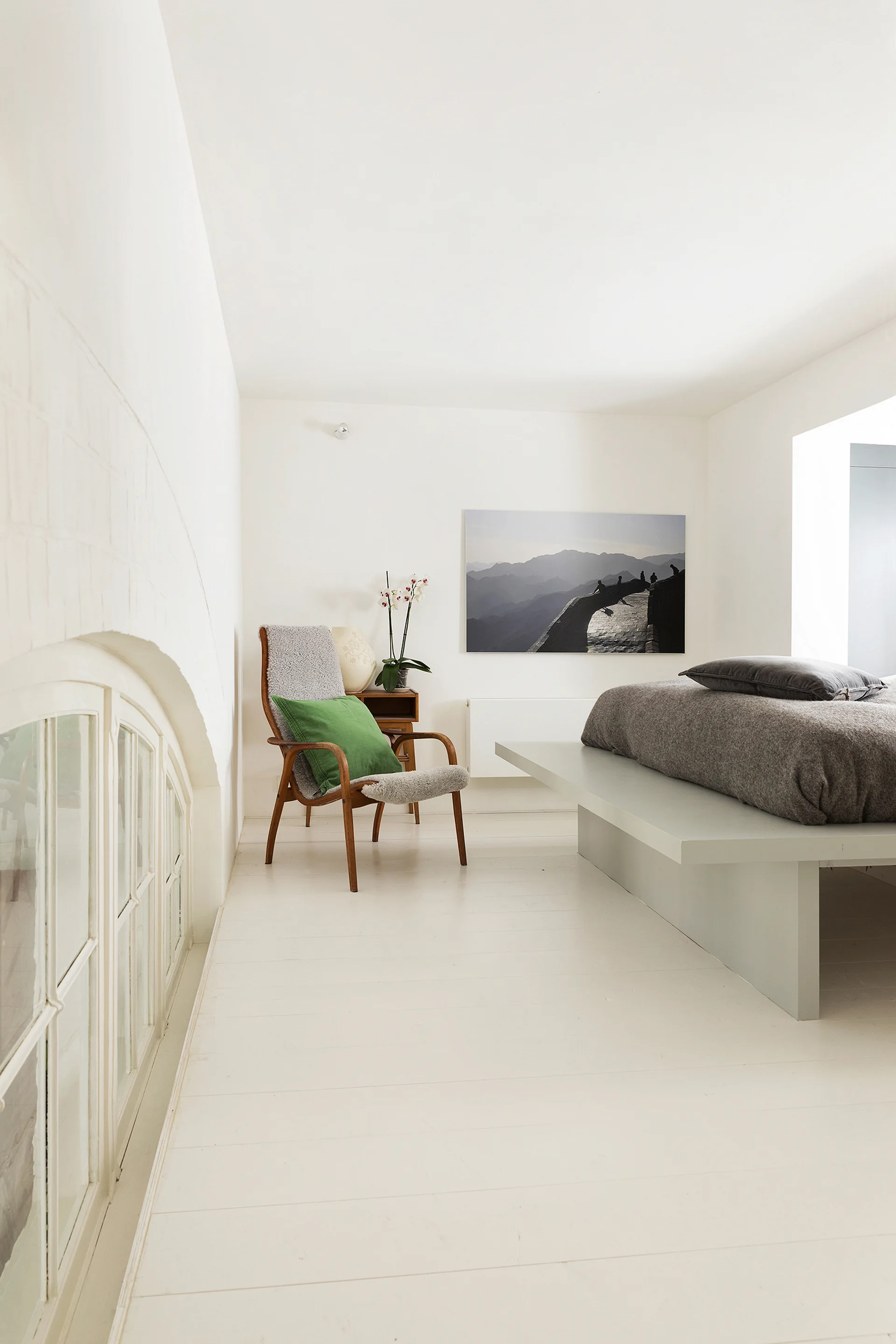 A minimalist bedroom with painted white wood floors, a low grey platform bed, a wooden armchair with a green pillow, and an arched window.